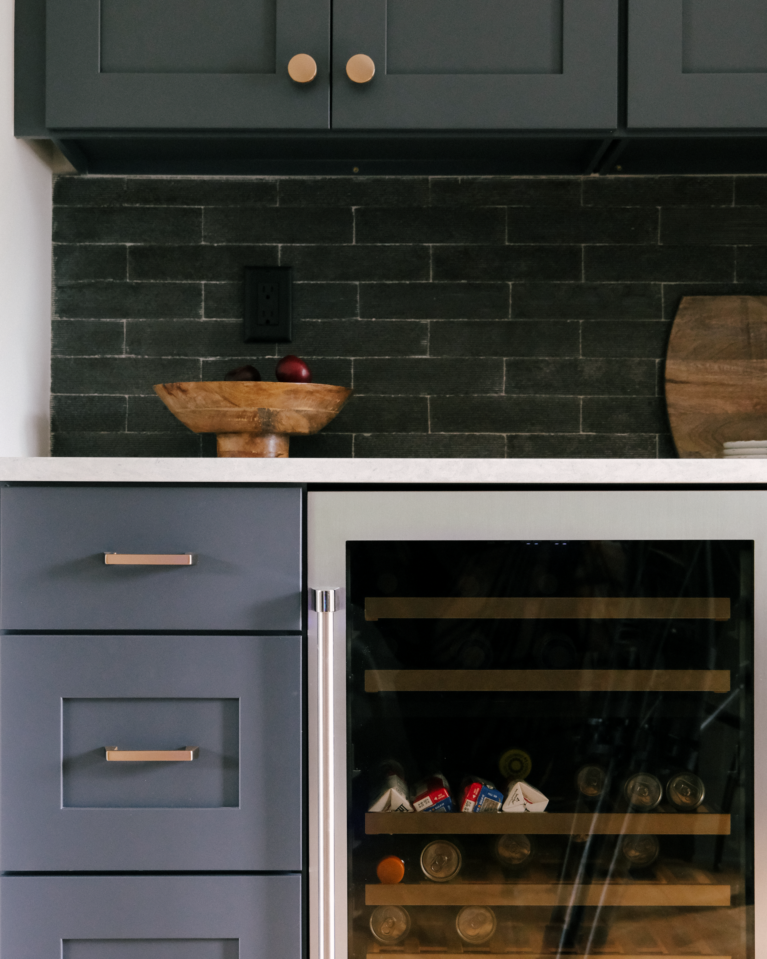 Moody light and dark blue kitchen cabinets and drawers with gold handles, above a beverage fridge. A wooden bowl with fruit and a chopping board sit on the white countertop.