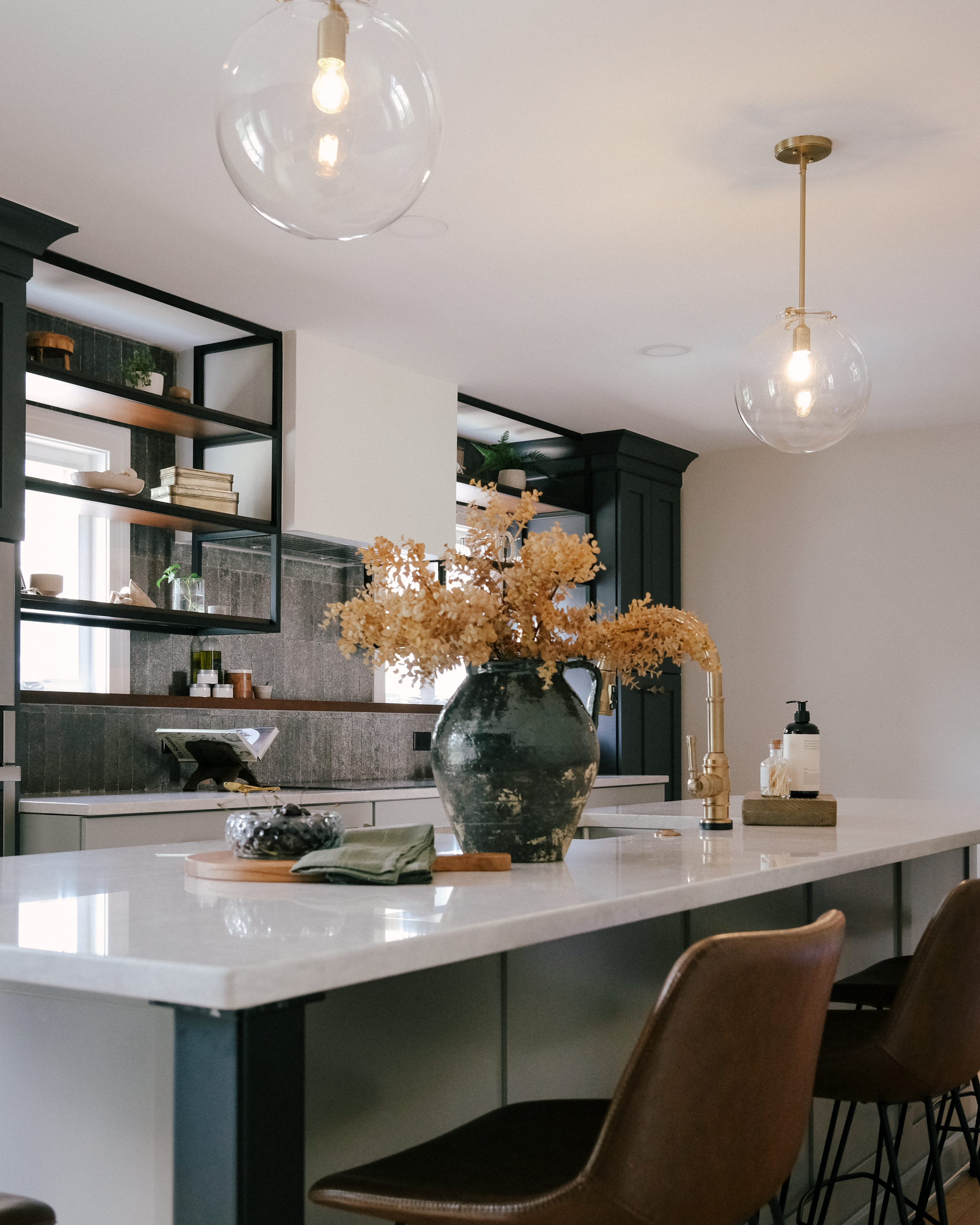 Modern kitchen with a white quartz island, brown leather chairs, and industrial open shelving over bright windows. A vase with dried flowers centers the counter, under two globe pendant lights. Calm ambiance.