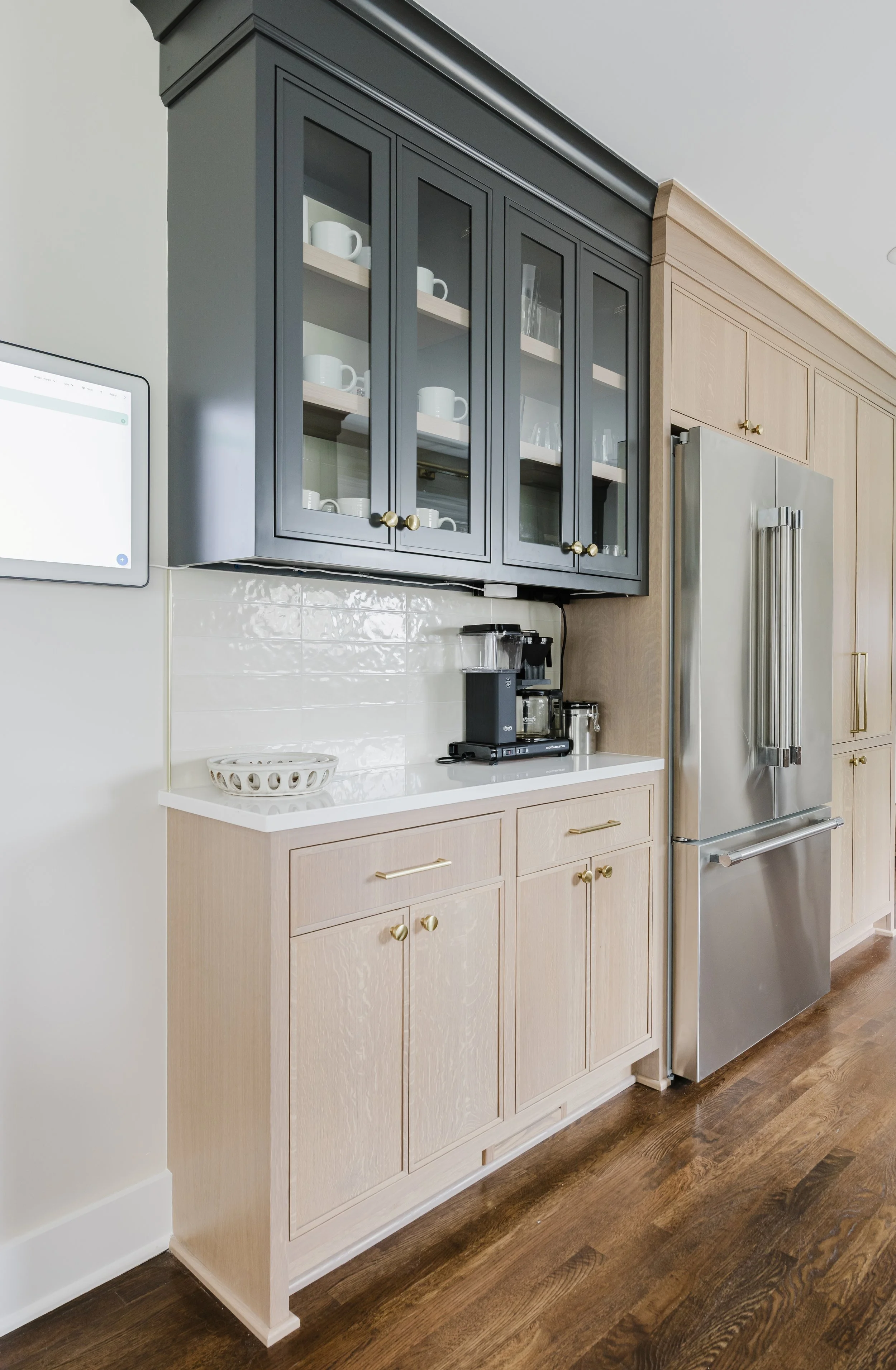 Modern kitchen corner with dark upper cabinets, glass doors showing cups, White Oak lower cabinets, a coffee maker, and a stainless steel fridge.