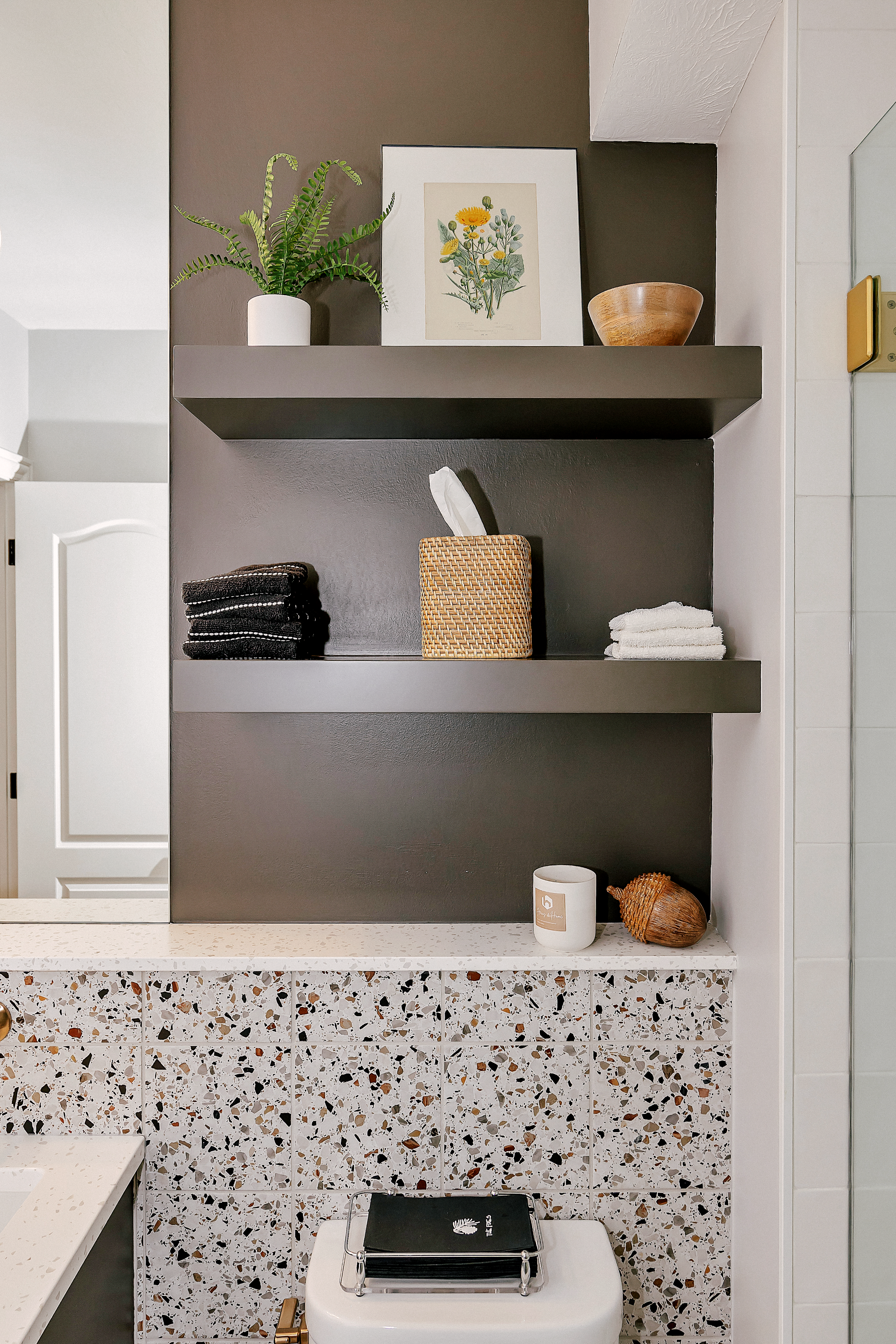 Modern bathroom with dark integrated shelves displaying a potted plant, floral artwork, bowls, and a woven box. A mosaic tile wall adds texture.