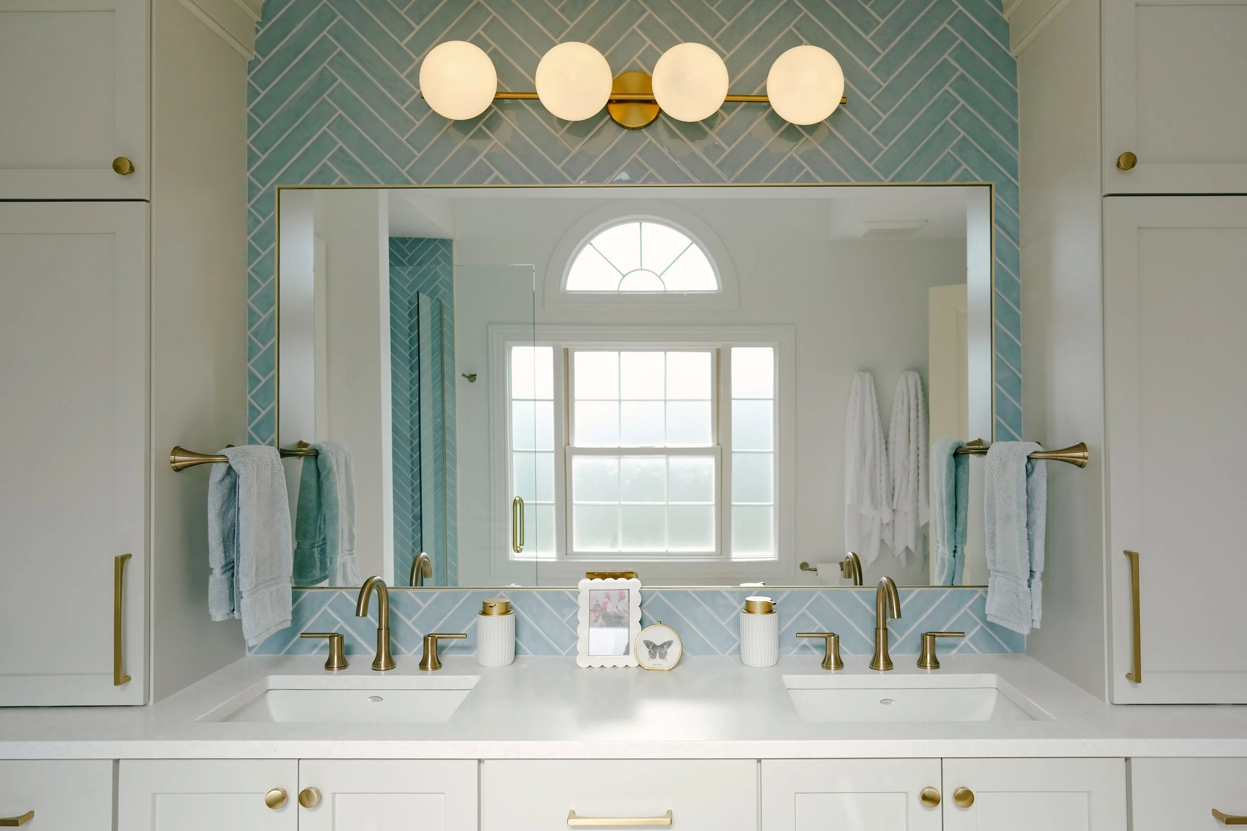 Elegant, bright children's bathroom with dual sinks, honey bronze accents, fixtures and hardware, and a large mirror. Beautiful blue herringbone tile backsplash and round vanity lights add a soft, playful touch.