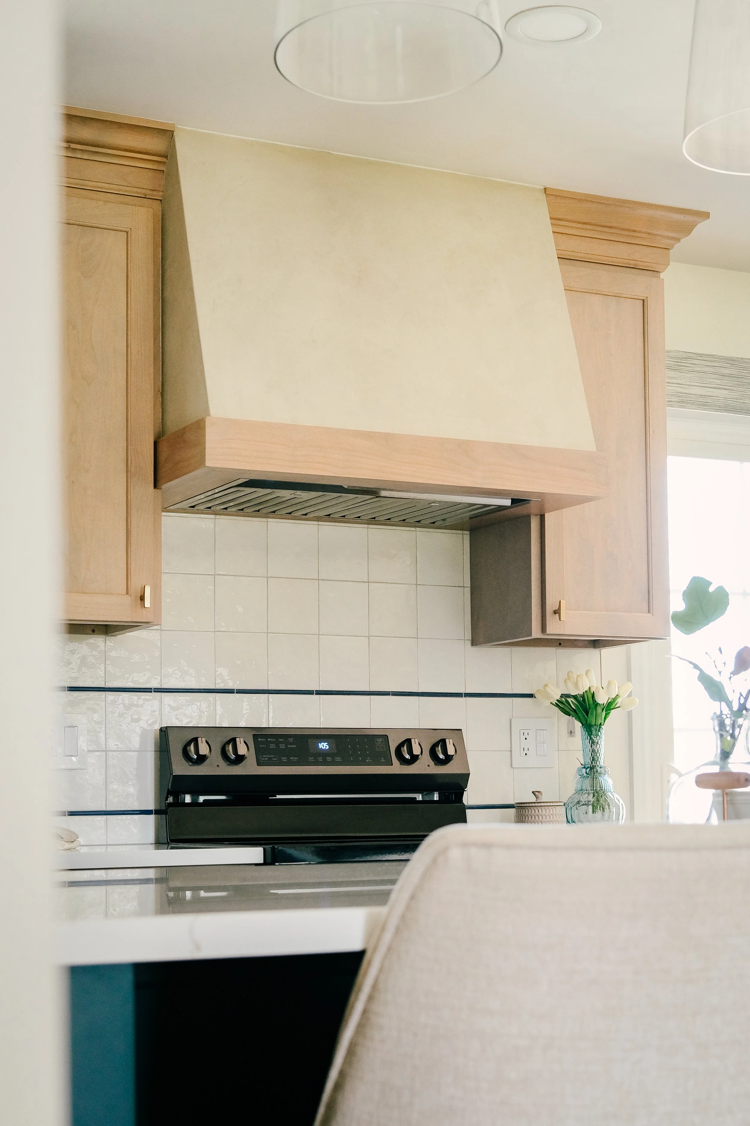 Charming, neutral kitchen with light wood cabinets and plaster range hood, black stove, white tiled backsplash, and a vase of yellow flowers on the counter.