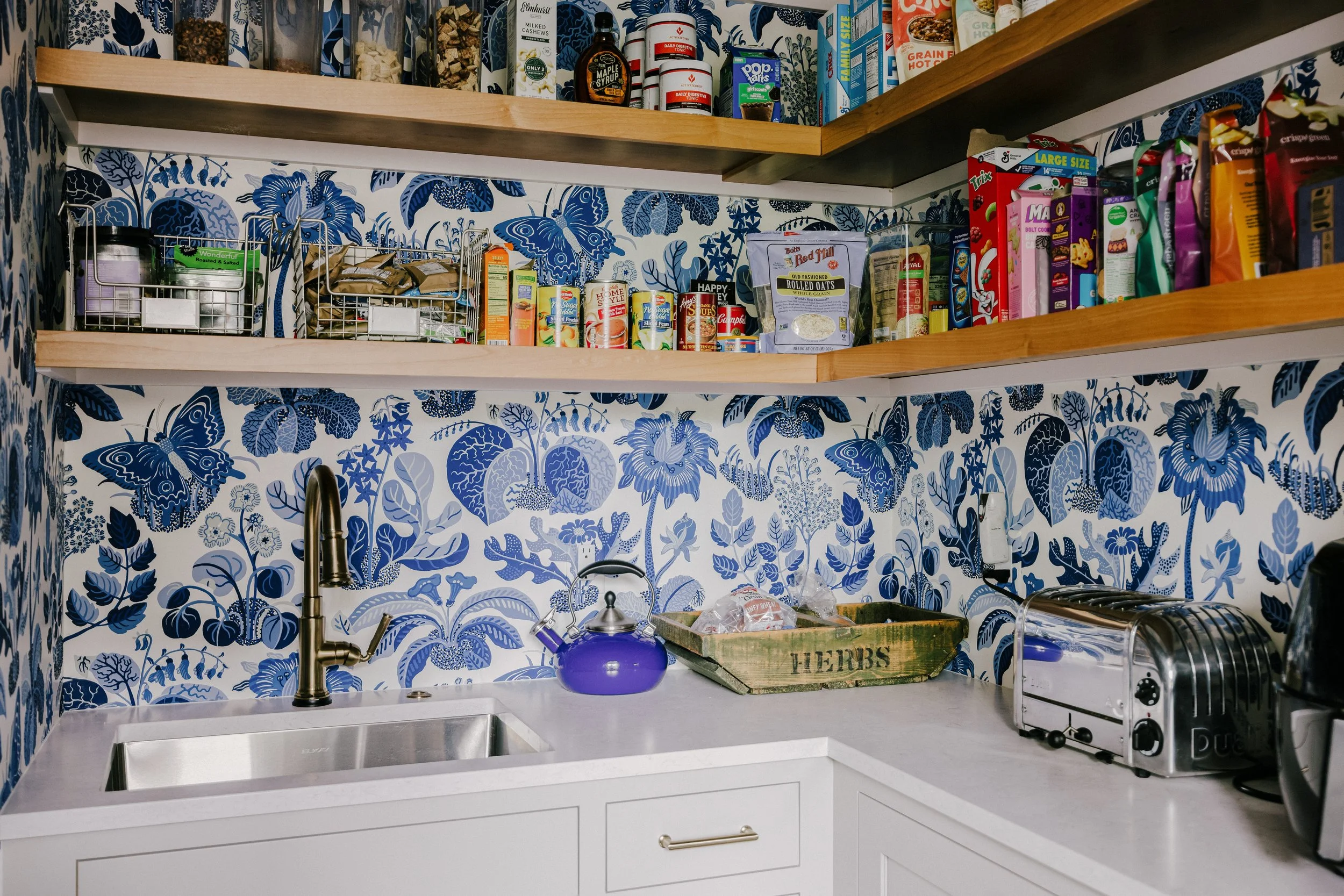 Quaint pantry with blue floral wallpaper, wooden shelves stocked with jars and cans, a small Elkay sink, gold Delta faucet, and a blue kettle. Cozy and organized atmosphere.