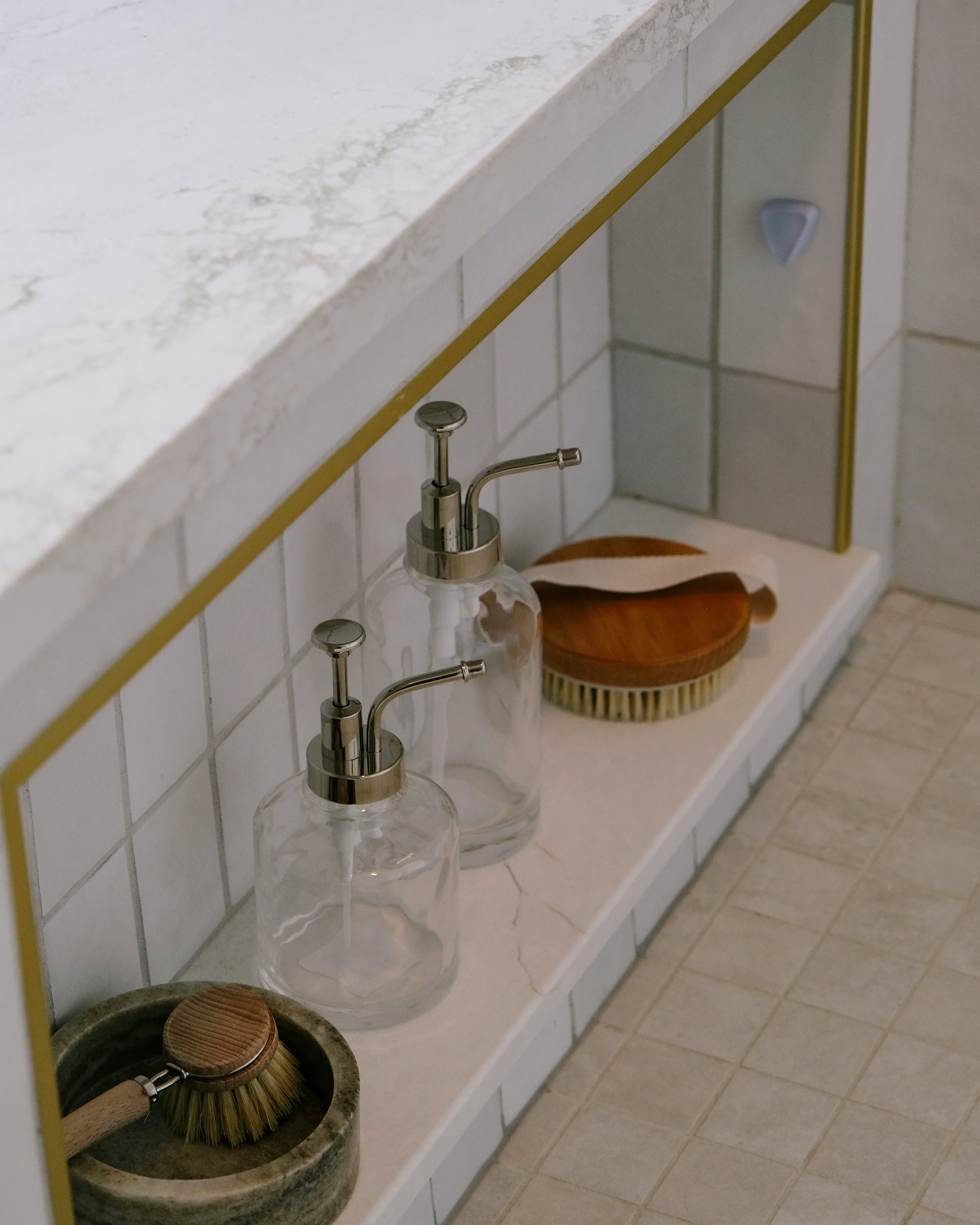 A Cambria quartz shower bench niche with gold accents holds glass soap dispensers and wooden brushes on a tiled ledge.
