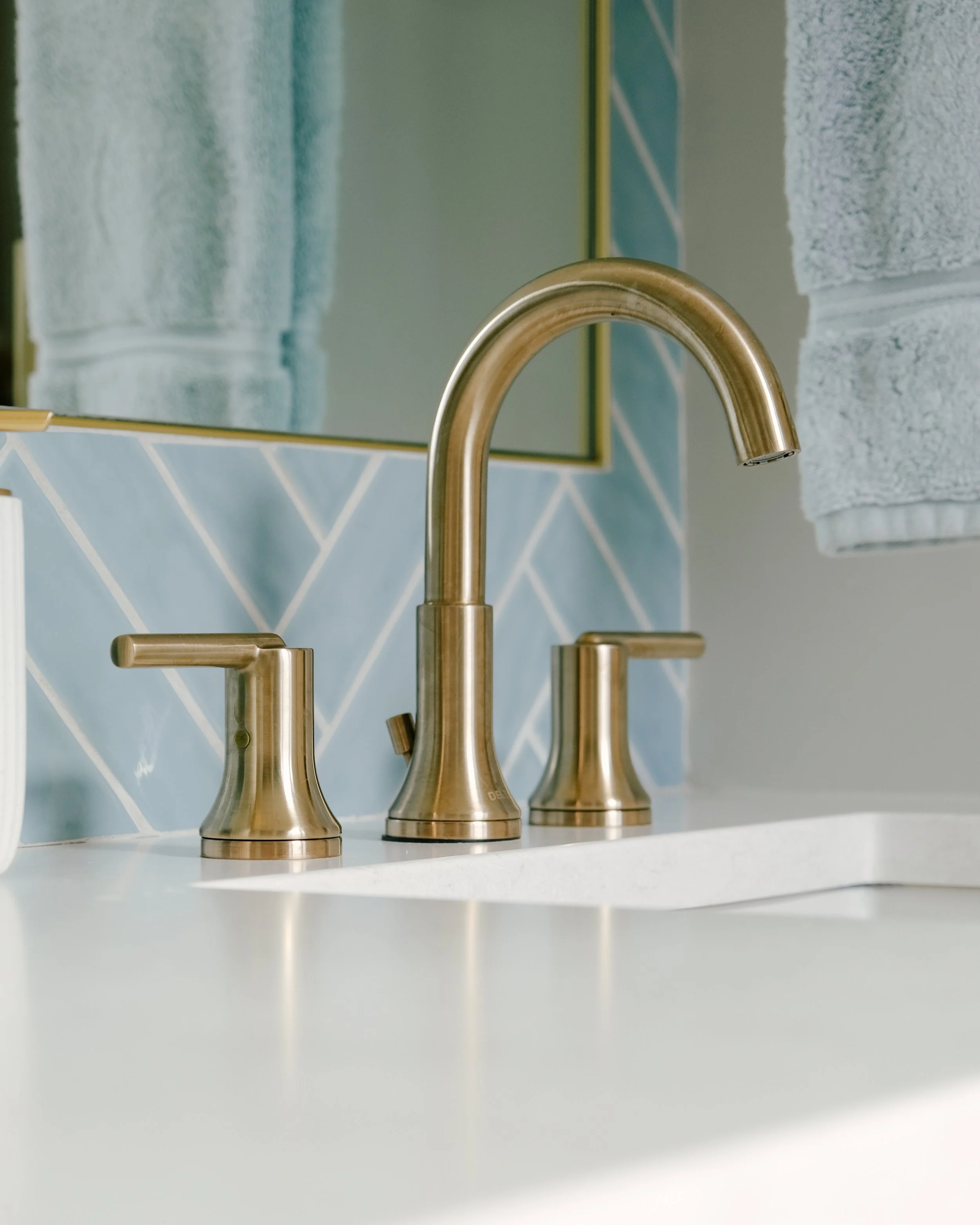 Close-up of a Delta Trinsic bathroom faucet with two handles on a white Cambria Seacourt countertop. A soft blue herringbone tile backsplash and a folded towel surround the fixture.