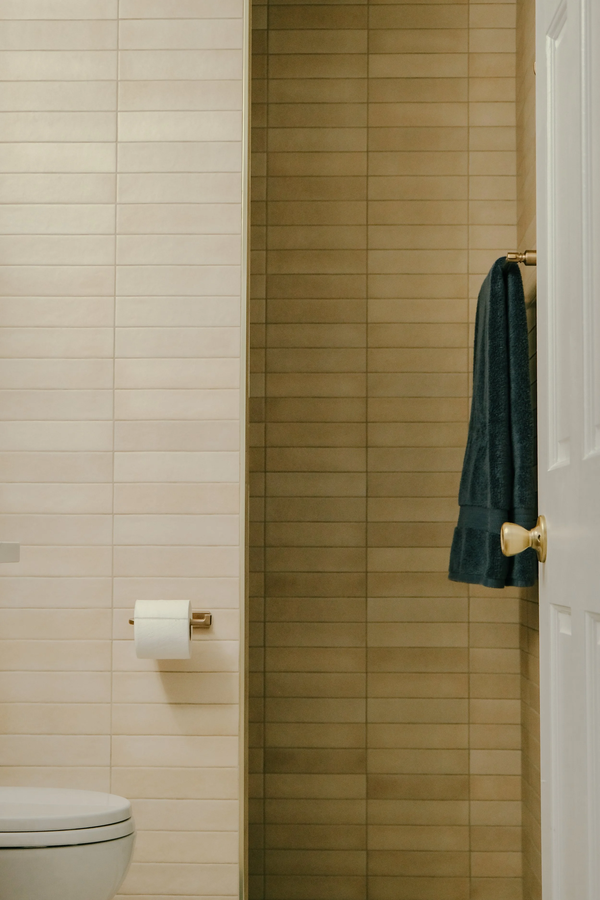 A minimalist bathroom features beige tiled walls leading to a shower, a white toilet, a roll of toilet paper, and a dark towel hanging on a door handle, conveying simplicity.