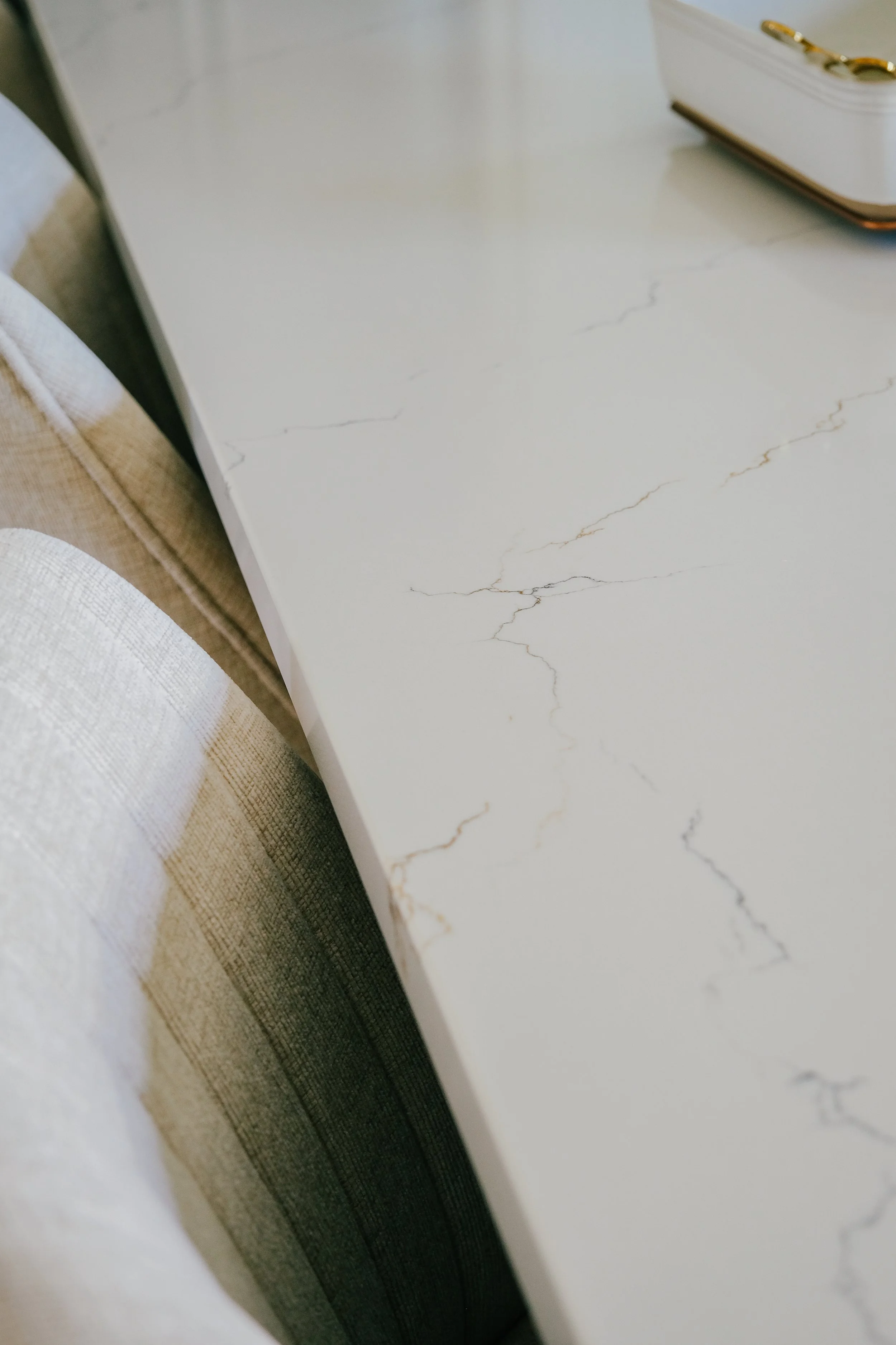 White Cambria quartz countertop with subtle gray and gold veining, next to a soft beige upholstered chair. A white box with gold accents is partially visible.