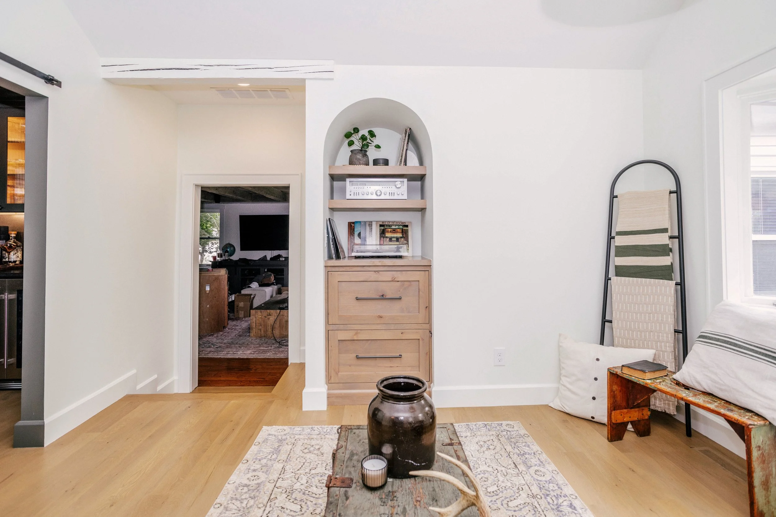 A cozy living space with light wood floors features an arched inset record player station with natural wood shelves above and cabinets below.