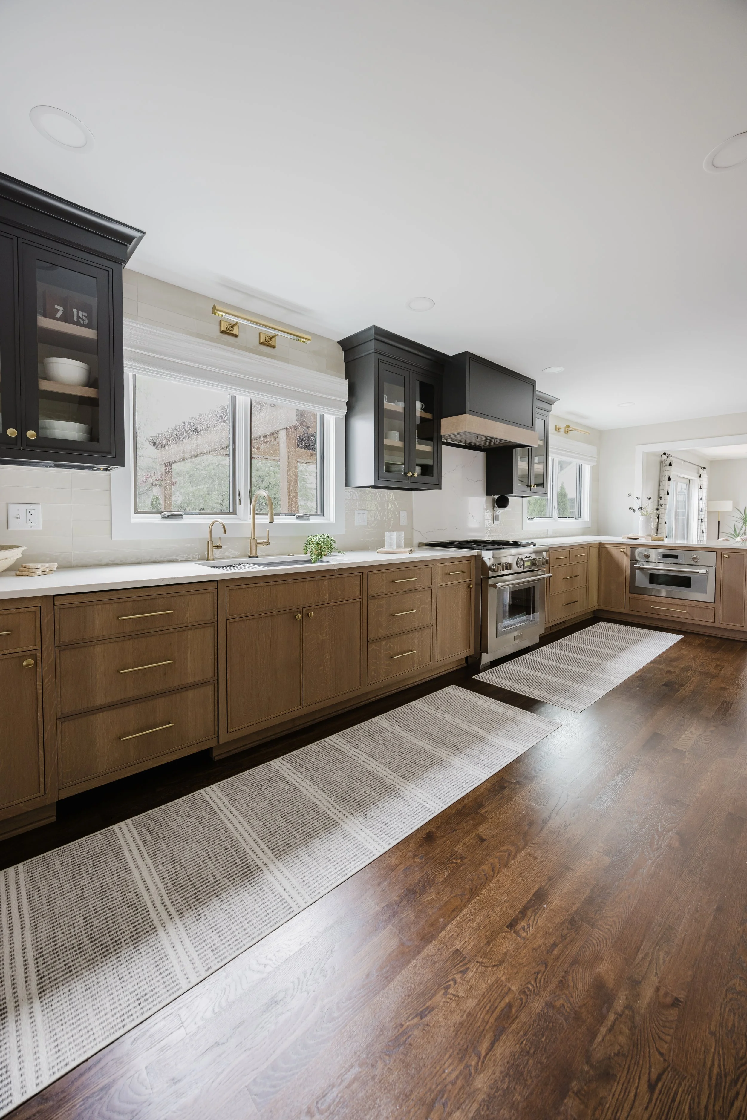 Spacious modern kitchen with dark wood floors, black upper cabinets with glass panels, and a stunning black and White Oak range hood. Natural light floods through large windows, creating a bright, inviting atmosphere.