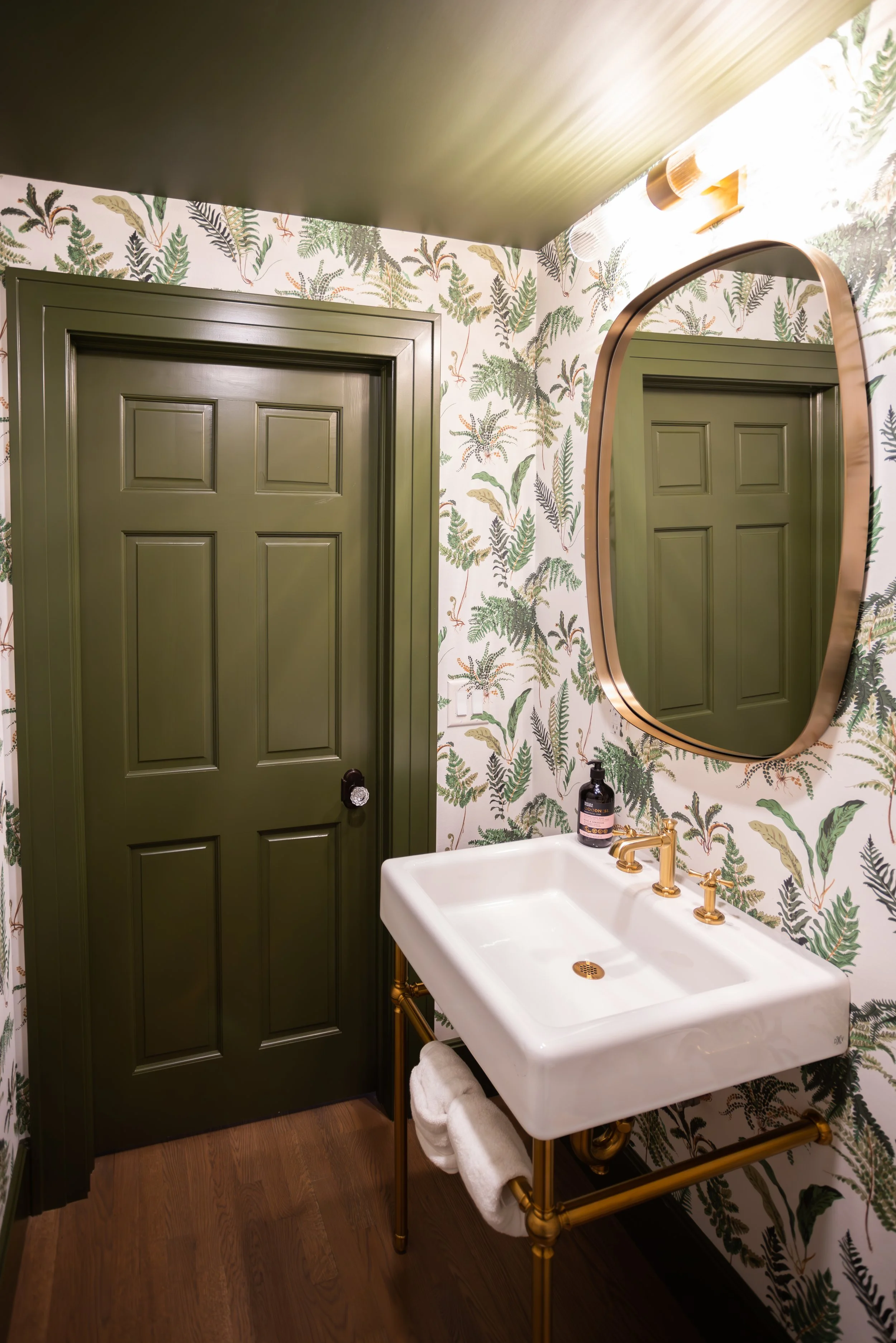 Bathroom featuring a moody green door and ceiling, fern-patterned wallpaper, and a gold-framed mirror. A white sink with golden fixtures and exposed piping and hardware adds a touch of elegance.