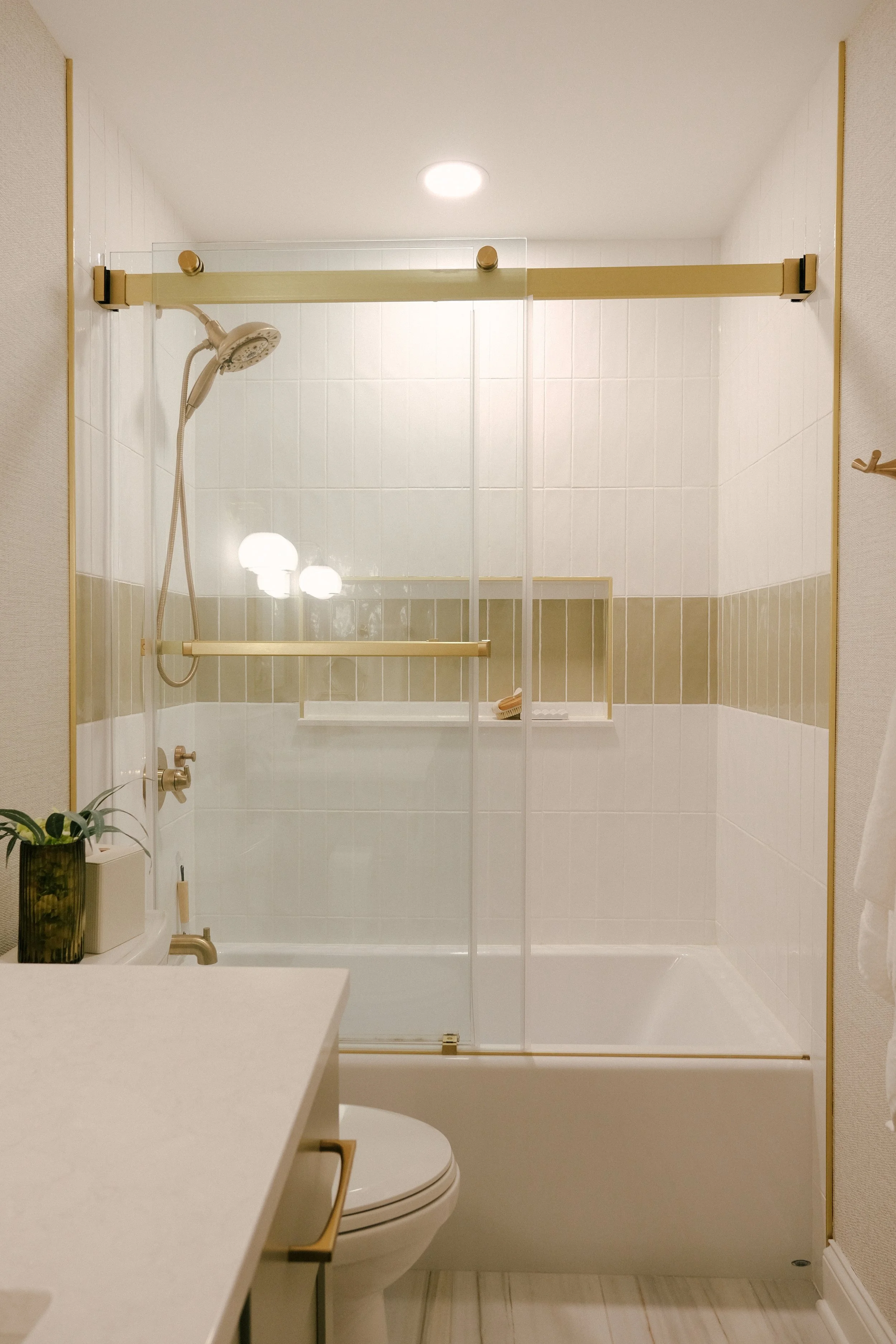 Modern bathroom with frameless sliding glass shower doors, gold accents, and beige tiles. A strip of green tile runs through the otherwise white shower. An integrated niche adds depth.