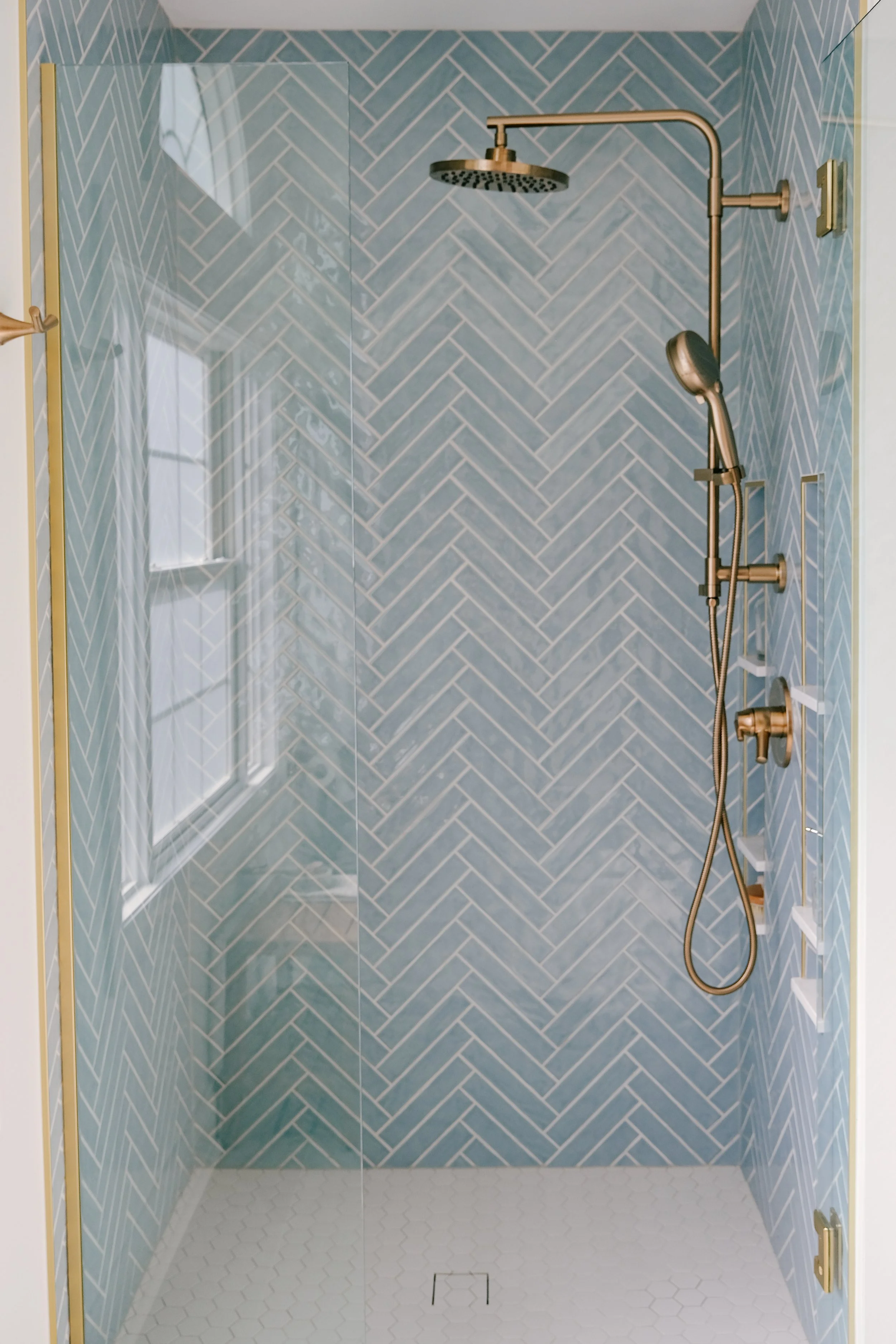 Timeless shower with blue herringbone tile, honey bronze fixtures, a large wall-mounted rain showerhead and smaller hand-shower, and a glass door. The space feels soft and serene.
