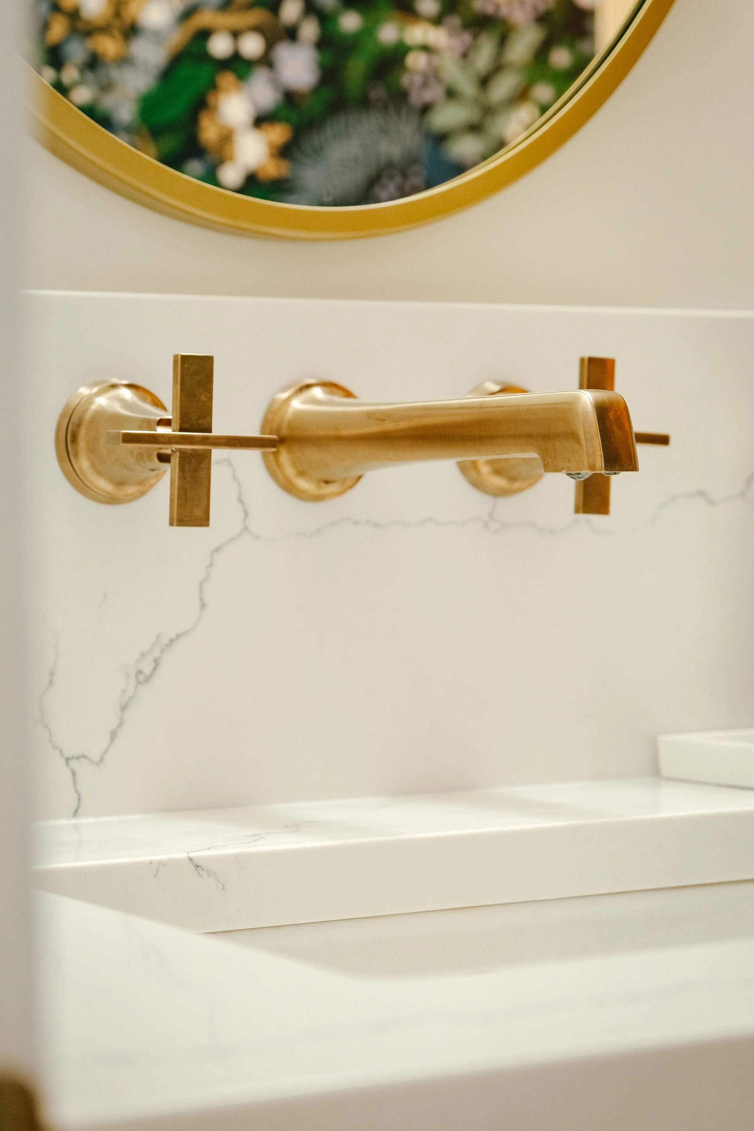 Elegant gold wall-mounted faucet set against a white quartz backsplash with subtle gray veining. A circular mirror with a gold rim reflects botanical wallpaper.