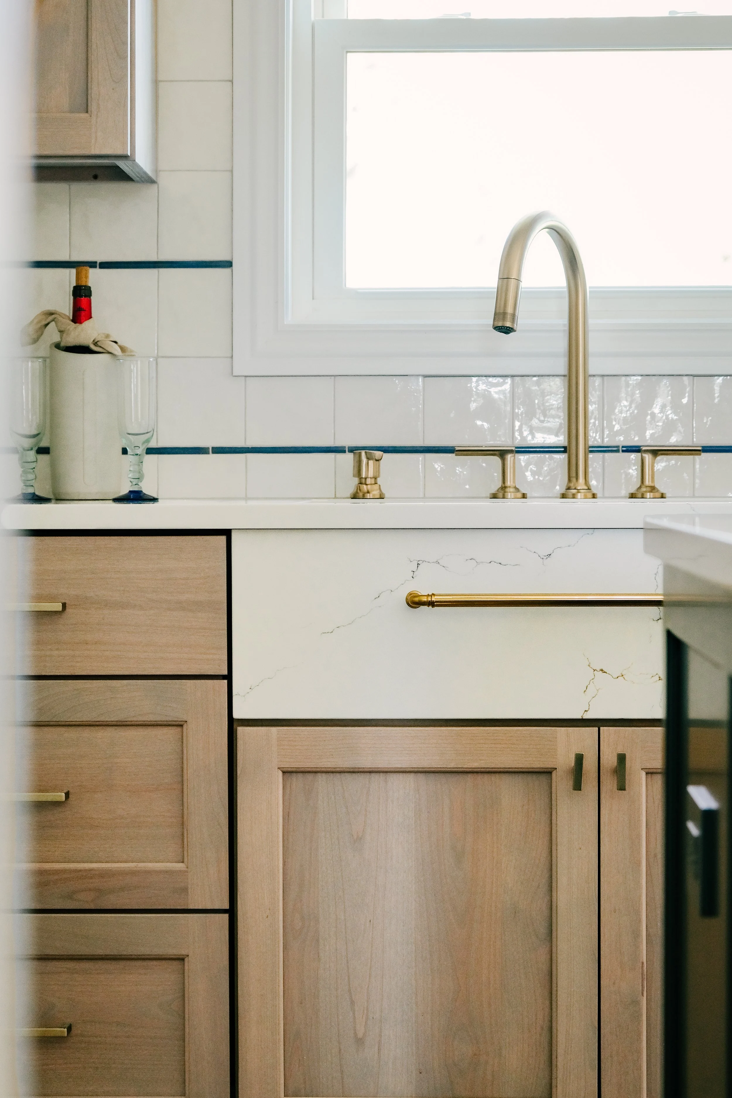 Neutral, quaint kitchen with custom stain wooden cabinets, a quartz countertop, and a bronze faucet beneath a window. A chilled wine bottle sits beside two glasses.
