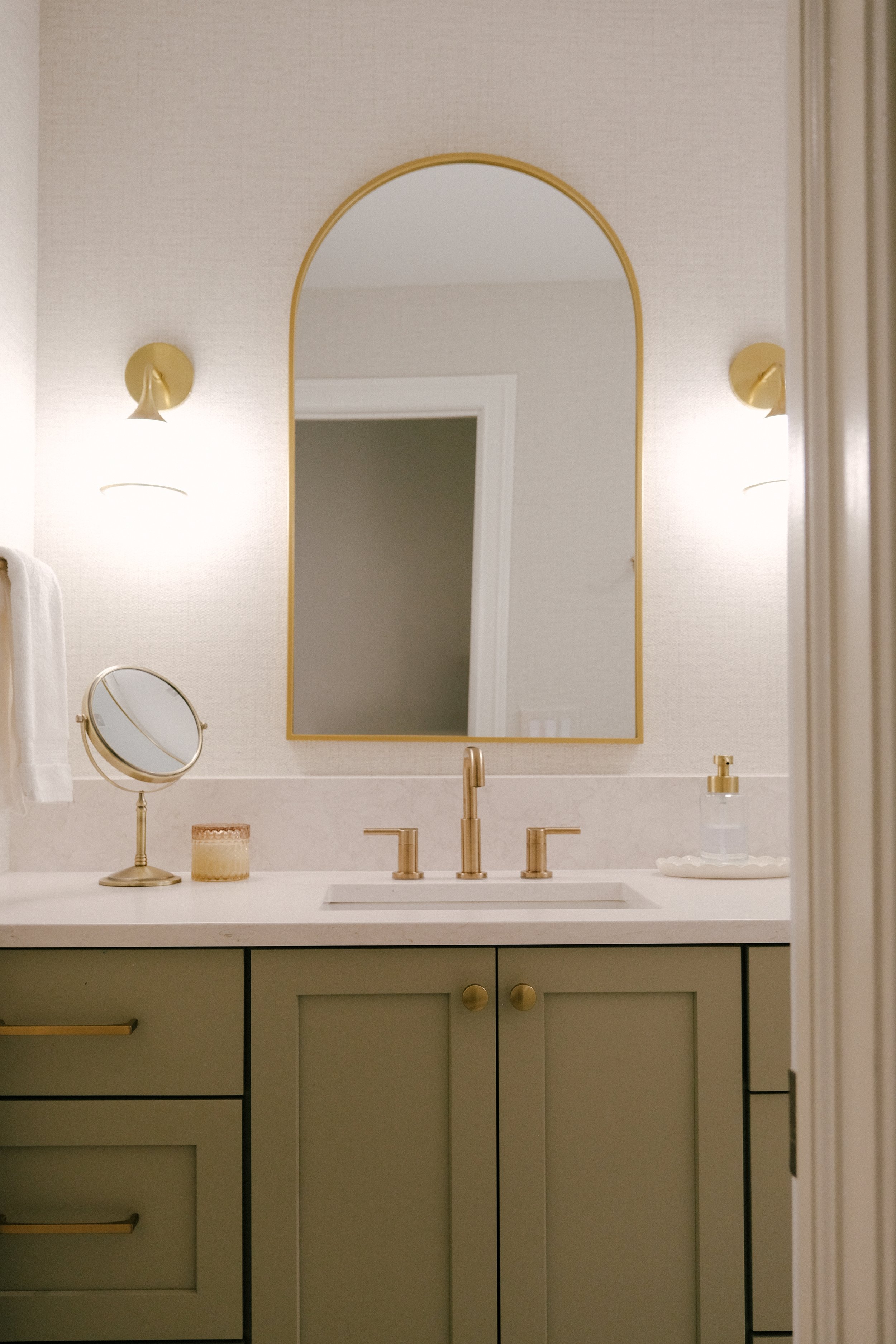 Elegant bathroom with slate green custom cabinets and a white quartz countertop. Gold faucet and hardware, arched mirror, and two wall sconces create a warm ambiance.