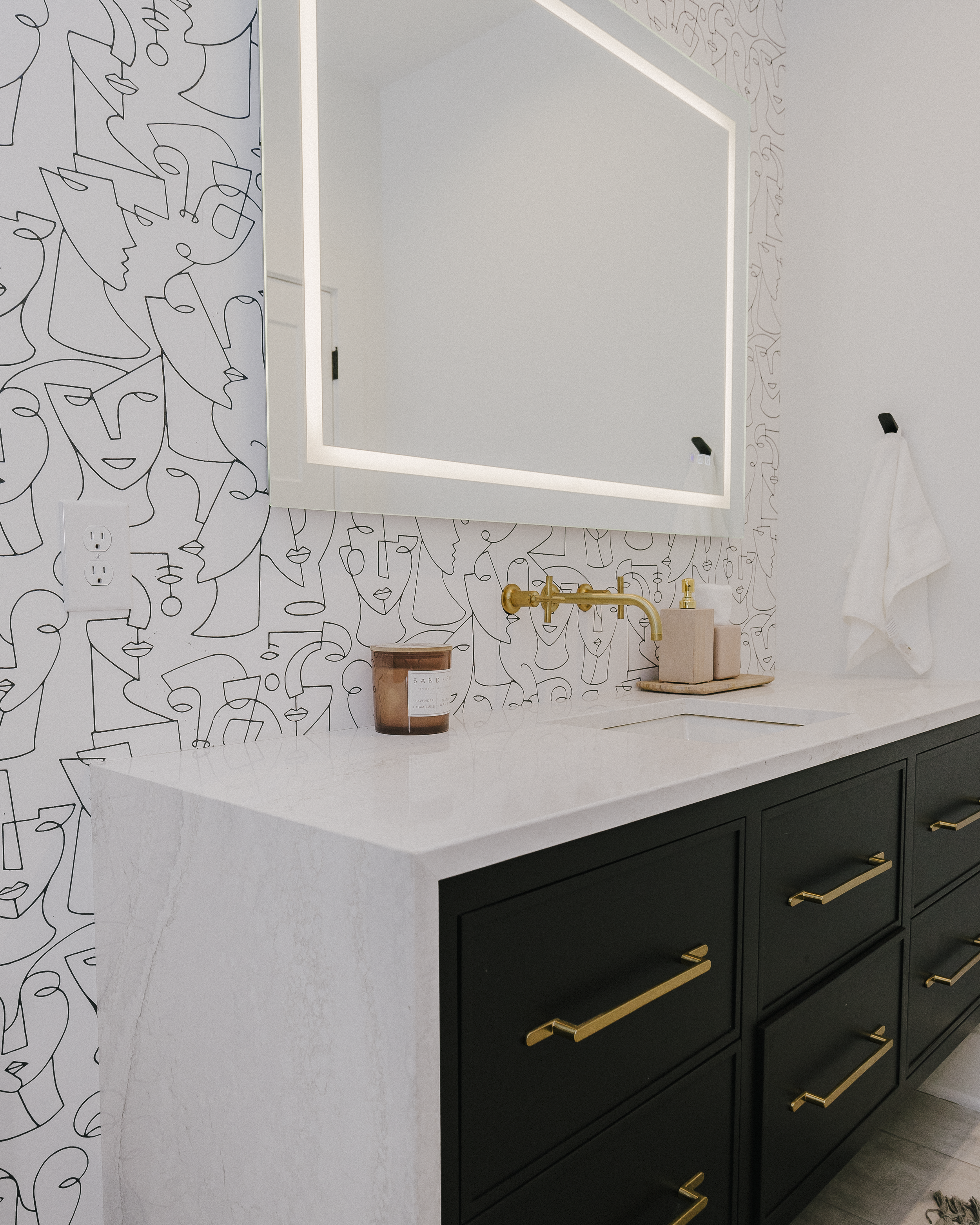 Modern bathroom with black vanity and waterfall-edge Cambria countertops, gold accents, and an LED illuminated mirror. Geometric line art wallpaper decorates the wall.