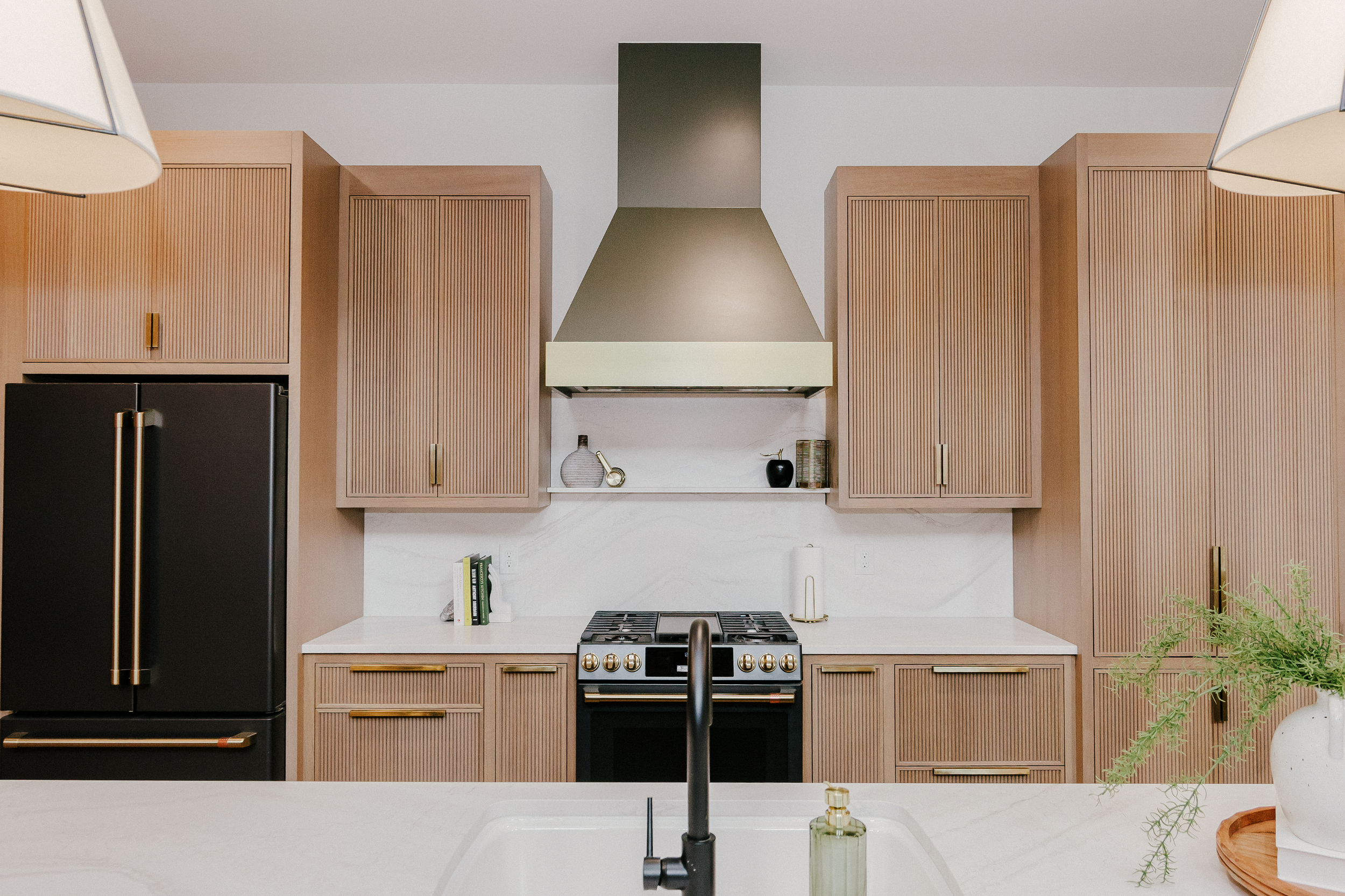 Modern, neutral kitchen with custom reeded cabinets, matte black fridge, and complimenting range. A sleek metal range hood with a gold band accent is centered. Greenery and warm lighting add a cozy touch.
