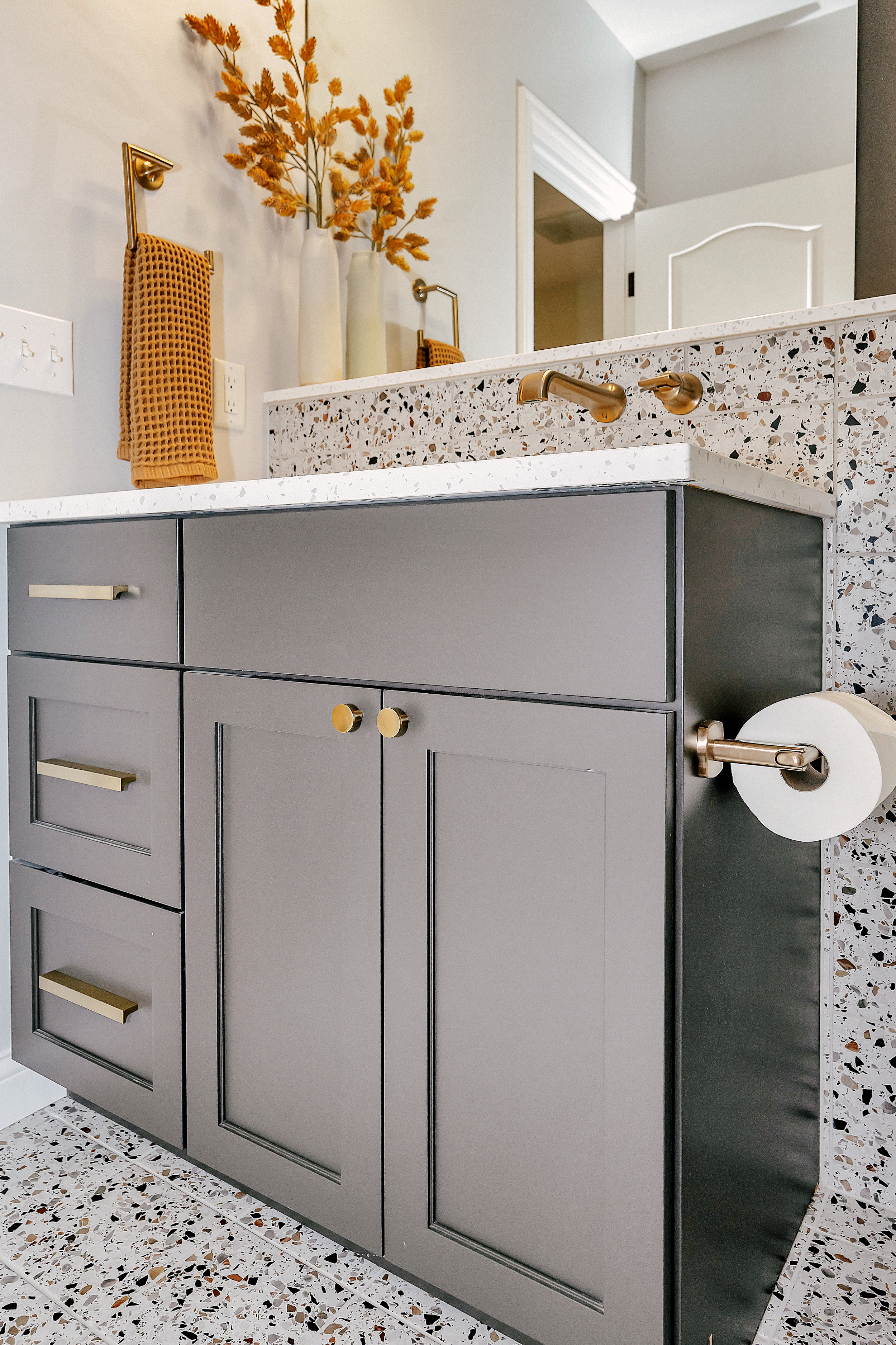 Modern bathroom with gray custom cabinetry, and floor-to-sink mosaic tile. Decor includes orange dried flowers and a mustard towel, creating a warm, stylish atmosphere.