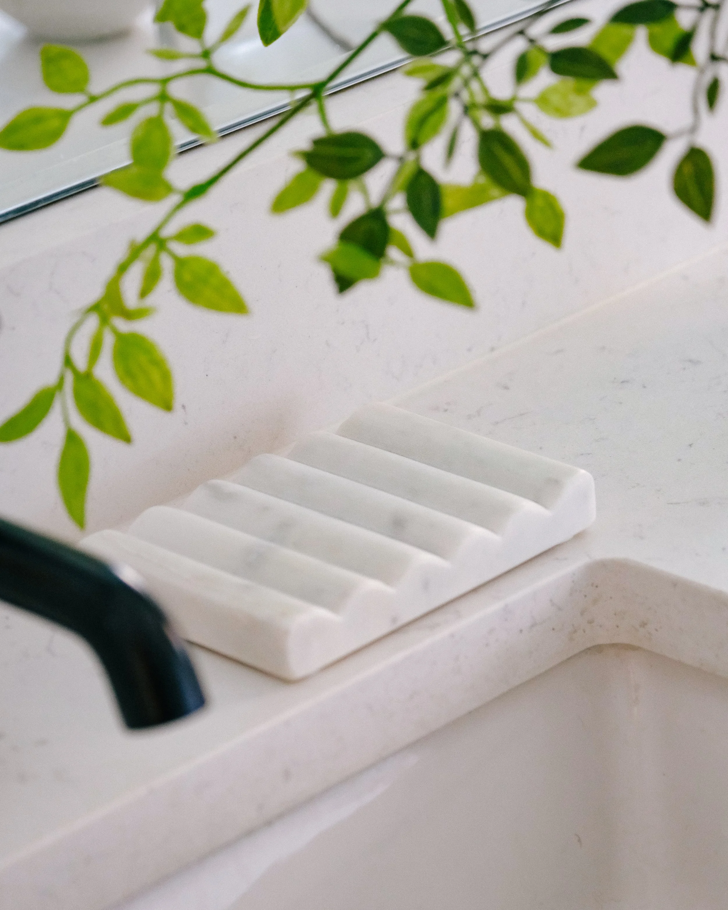 Close-up of a minimalist white soap dish on a white quartz bathroom counter, with lush green leaves hanging above, creating a fresh, tranquil ambiance.