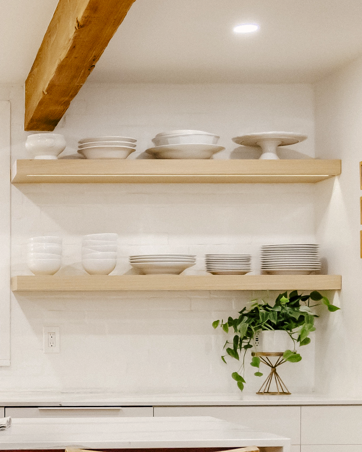 Open wooden kitchen shelves hold white dishware under an exposed beam. White painted brick acts as a backdrop for the kitchen scene.