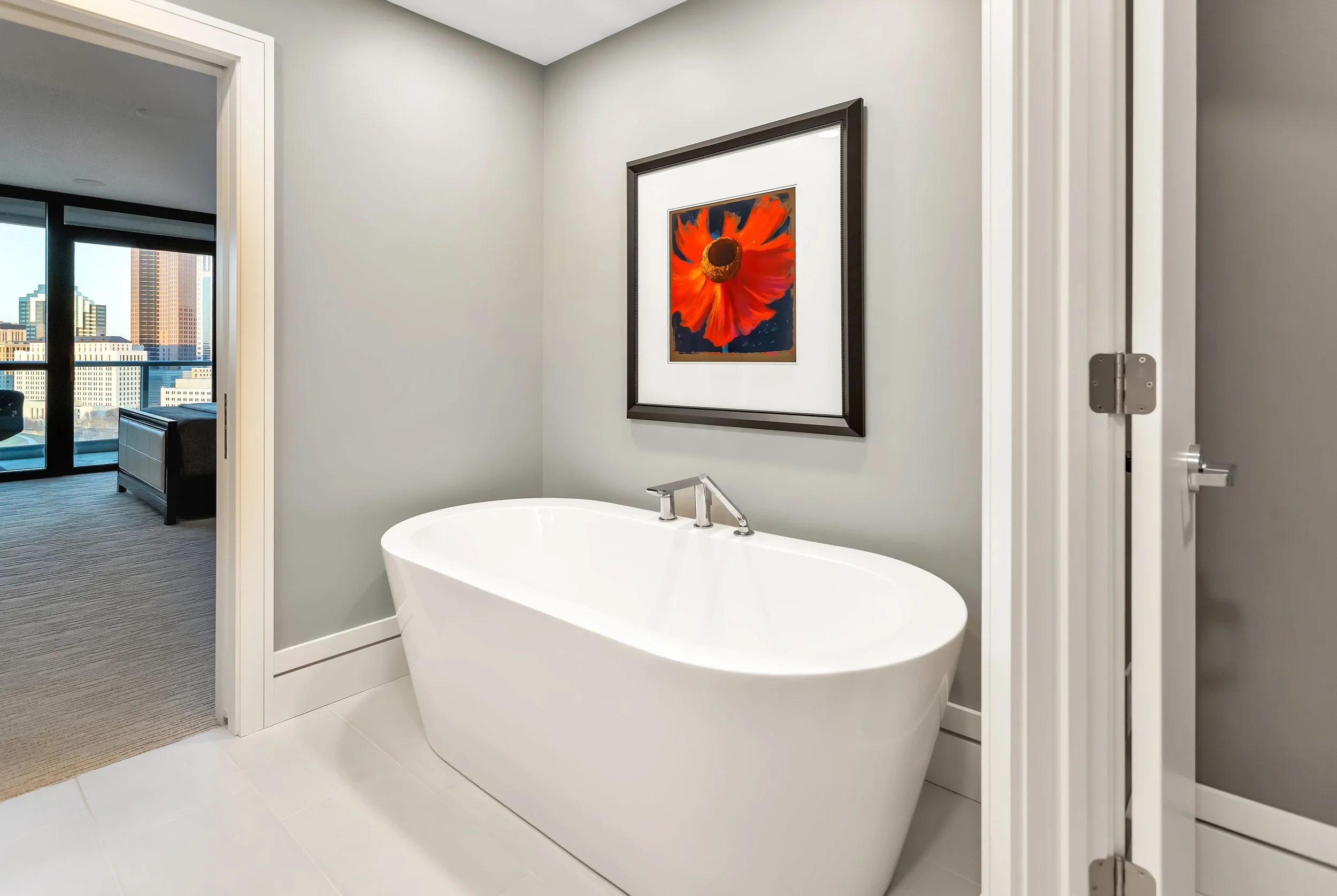 A modern bathroom with a sleek white bathtub and a vivid red photo on the wall. An open door reveals a bedroom with city views.
