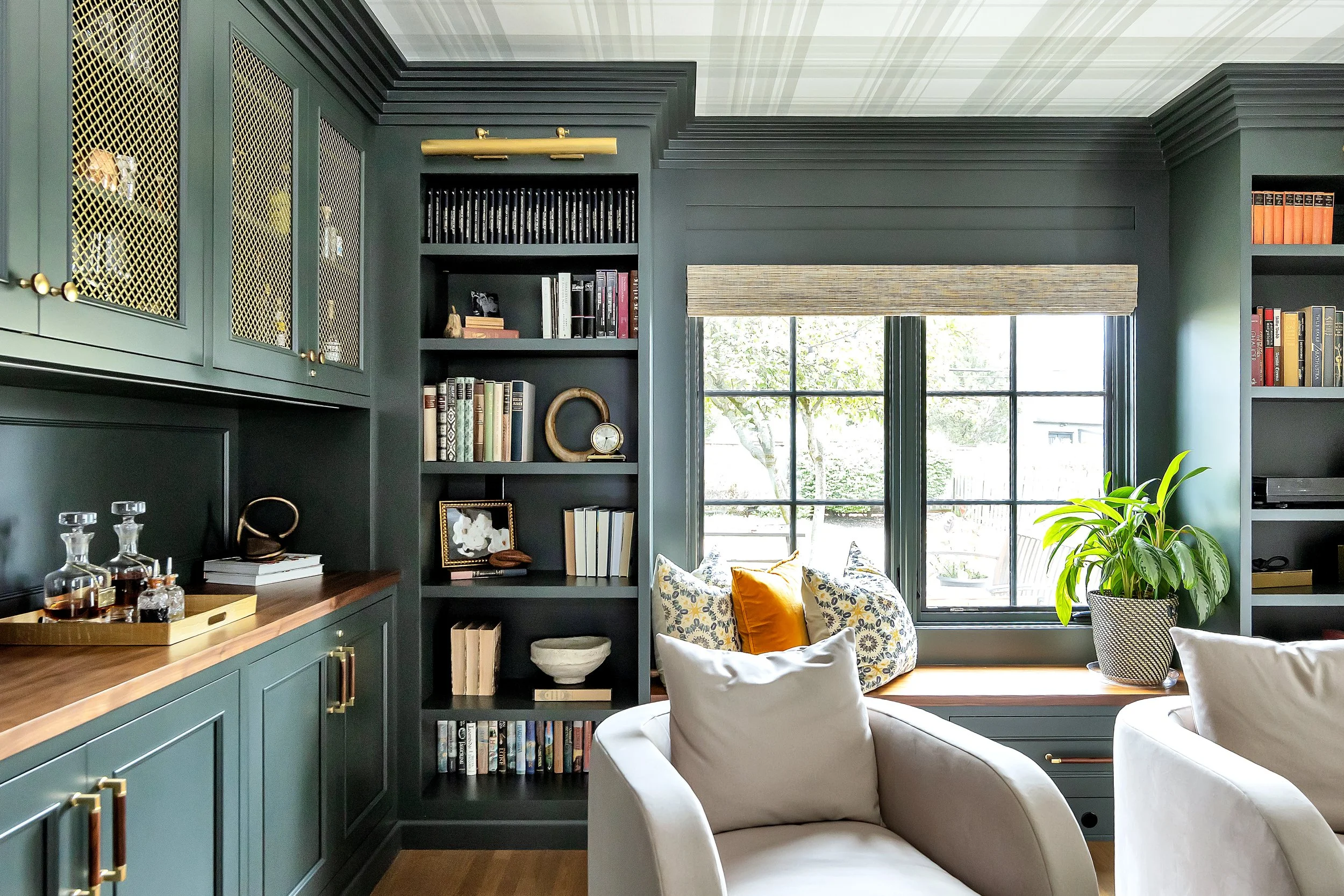 Cozy, contemporary library and dry bar with moody green shelves, books, and decor. Light streams through a window creating a tranquil setting.
