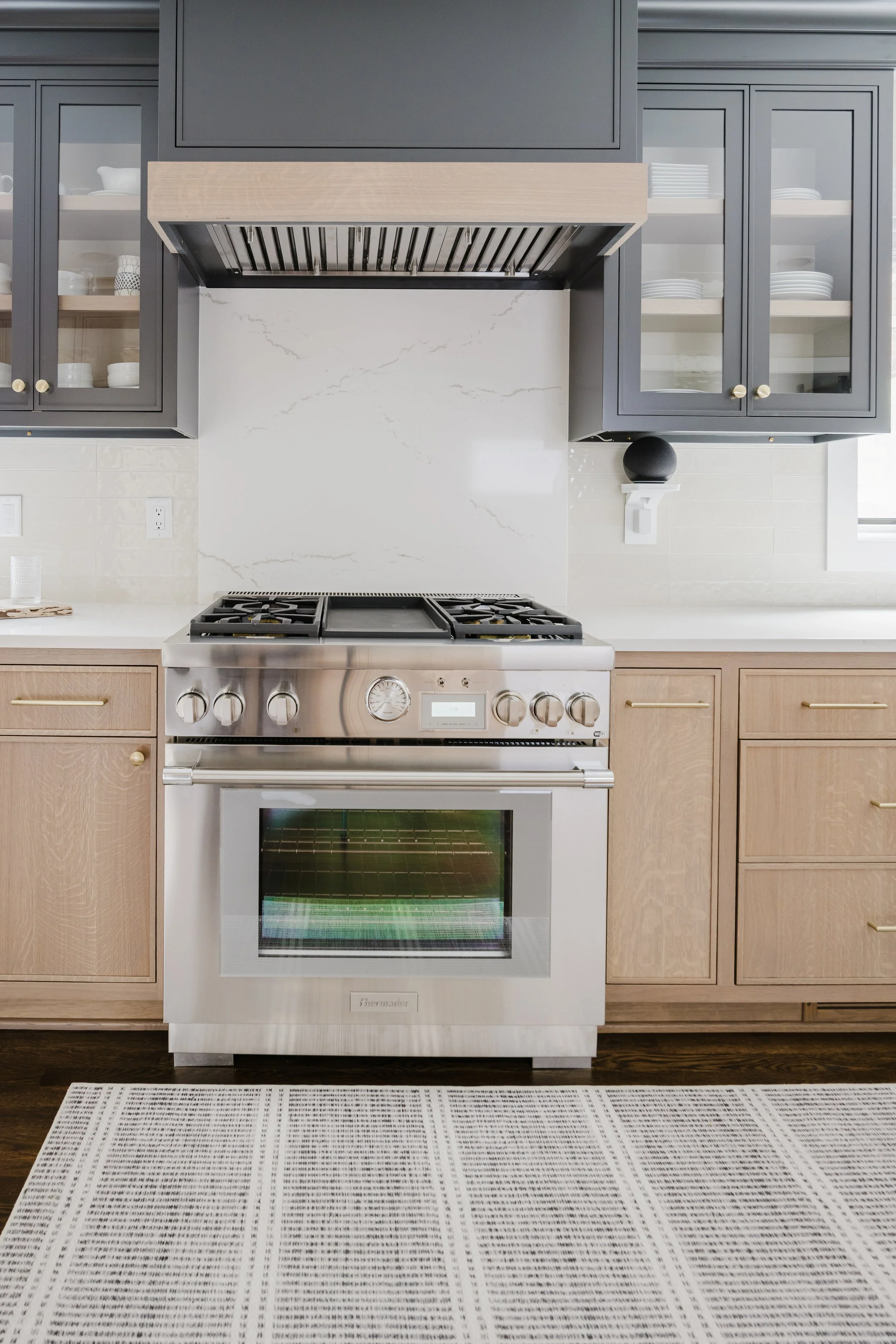 Modern kitchen with a gas Thermador range, flanked by light wood cabinets. Glass-door cabinets overhead show neatly stacked dishes. Elegant and tidy.
