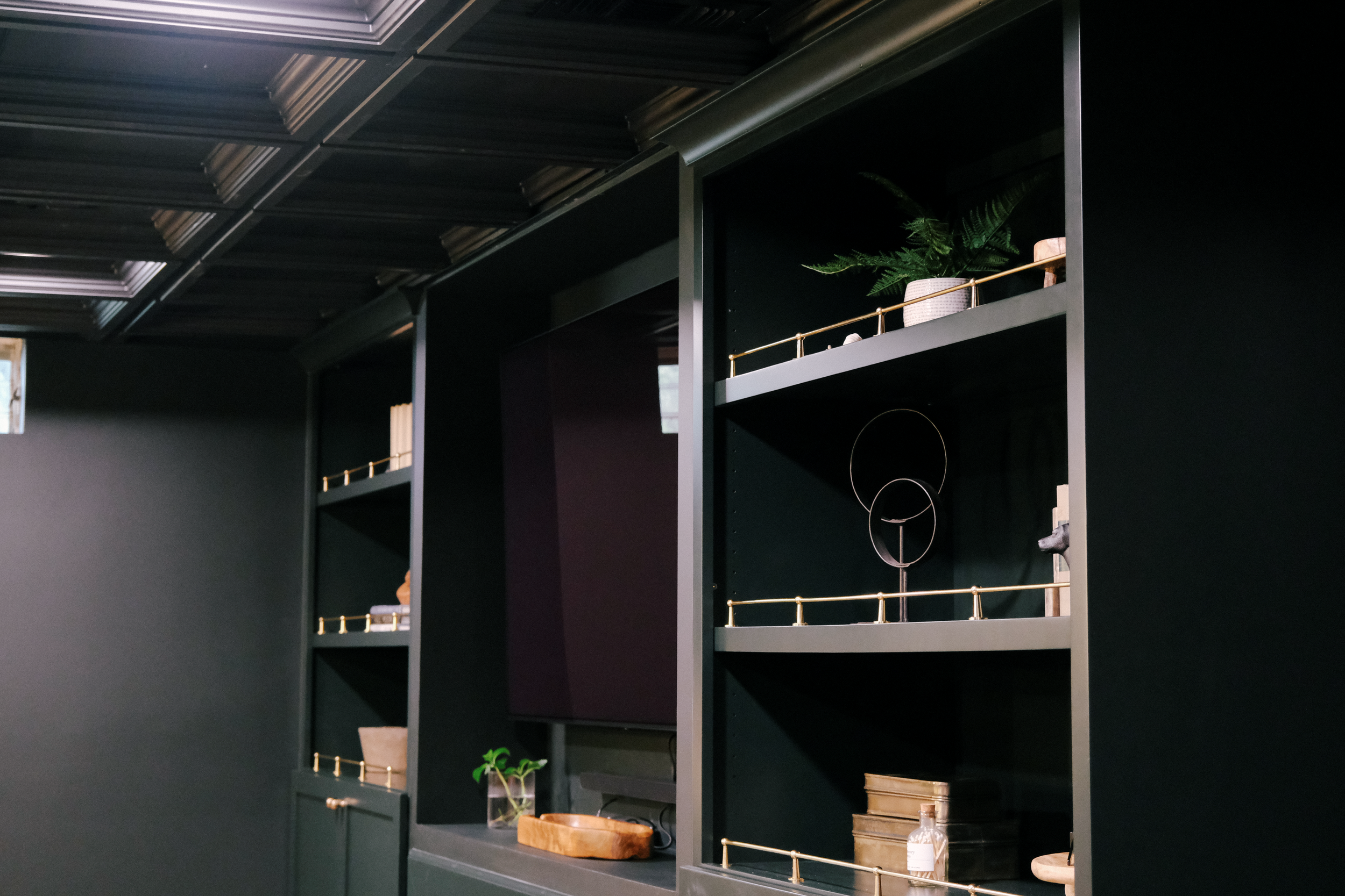 Moody color drenched basement with integrated shelves filled with decor items like potted plants, abstract art, and books. A TV sits in the middle of the shelving units. The room has a coffered ceiling, creating a cozy, sophisticated ambiance.