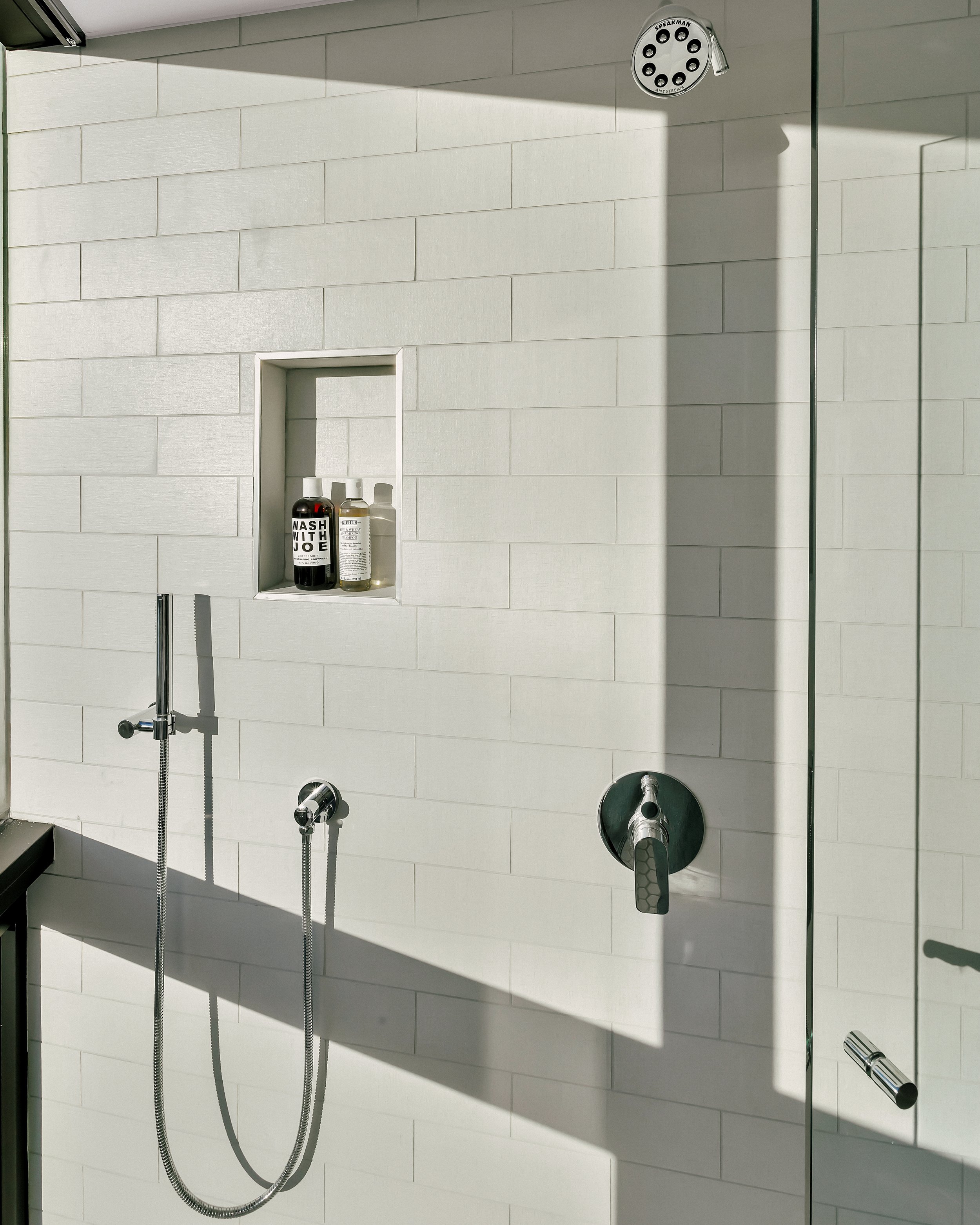 Modern shower with tile walls, featuring an inset niche. A sleek handheld shower and controls are visible, casting soft shadows.