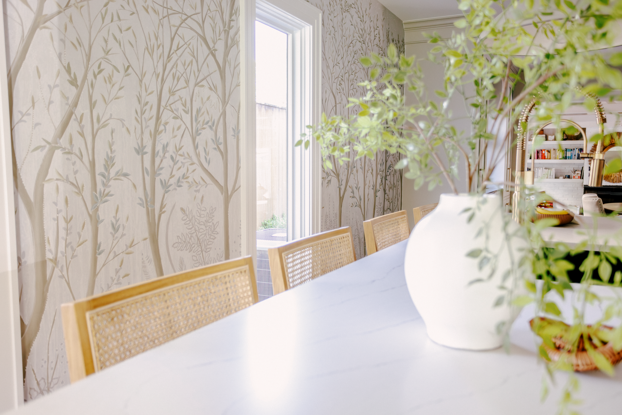 Neutral kitchen island with light wooden chairs, and a Cambria quartz top. Tree-patterned wallpaper mural adds a serene, natural touch.