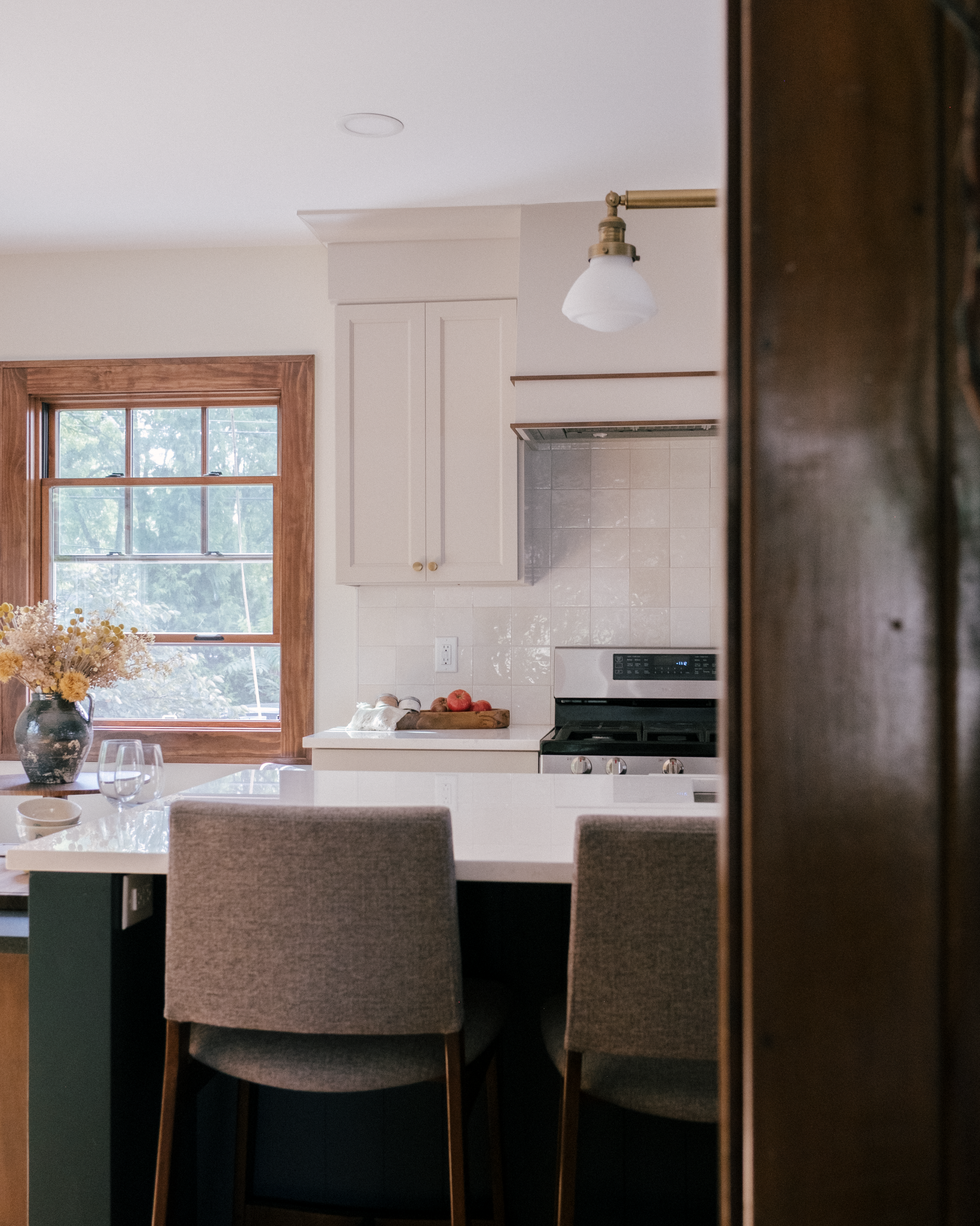 A serene kitchen with soft lighting, featuring a large blue island topped with Cambria quartz, a gorgeous and simple white hood with accent details, a vase of dried flowers, and a window offering a view of greenery outside.