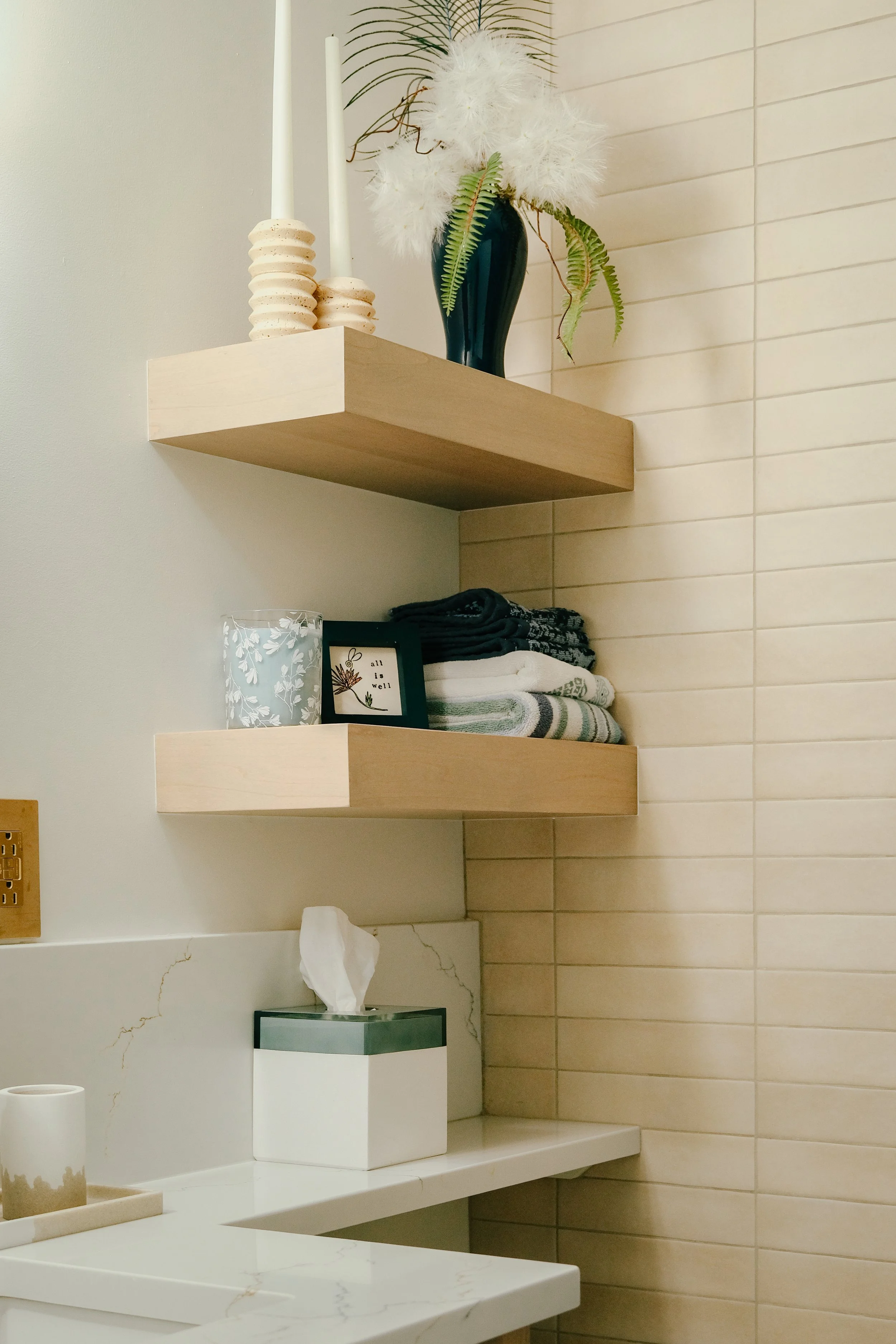 Modern bathroom with beige tiles and wooden built-in shelves. Top shelf has tall candles and a vase with pampas grass; lower shelf holds towels and a small framed picture.