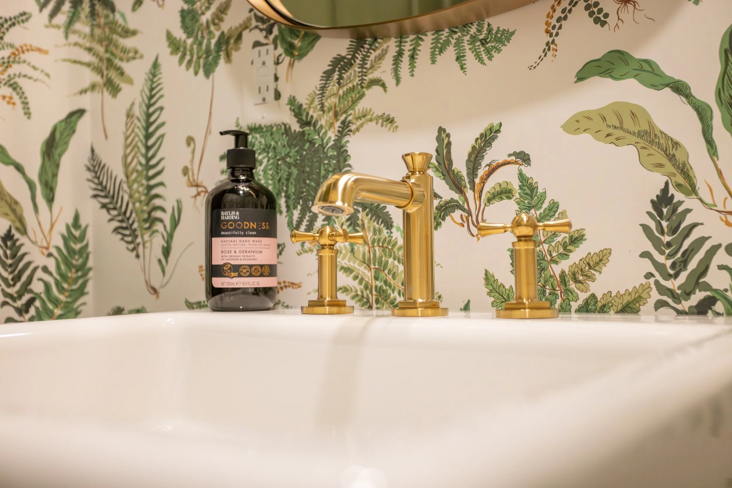 Stylish bathroom sink with elegant gold faucet and a soap dispenser, set against a lush green fern-patterned wallpaper, creating a fresh, quirky feel.