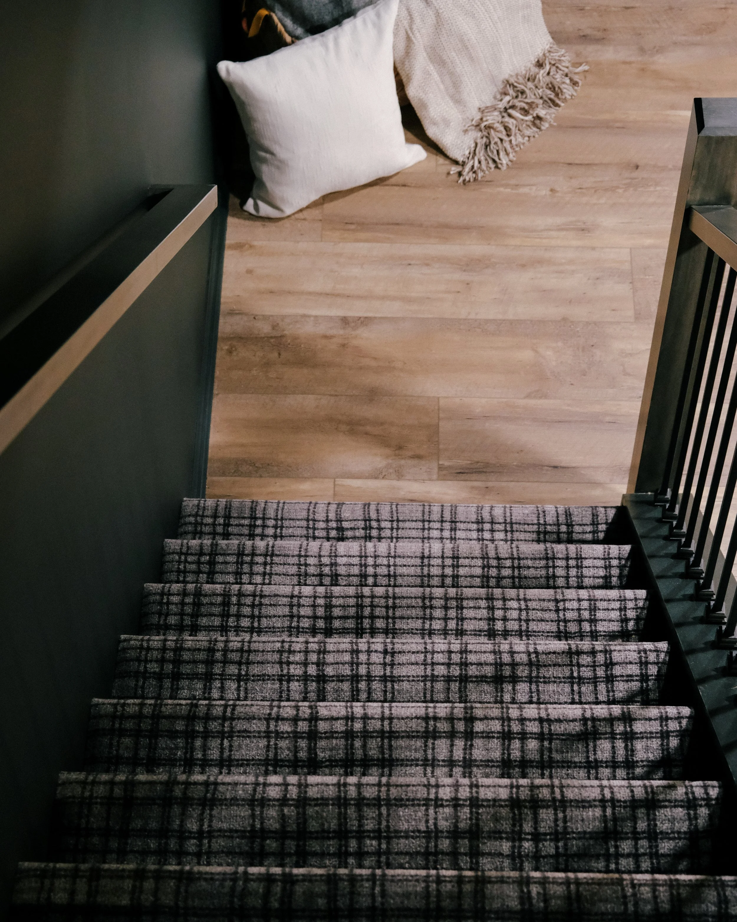 A staircase with plaid carpet leads down to a wooden floor. Two cozy pillows and a fringed blanket lie at the stair's base, creating a welcoming feel.