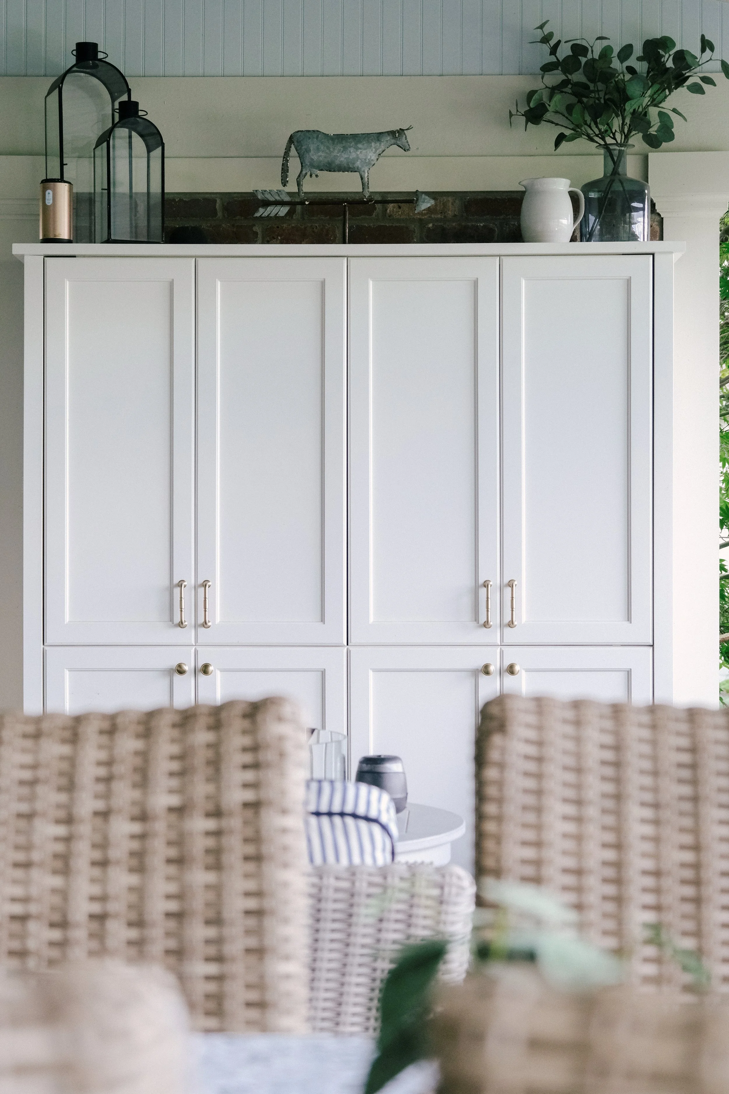 A white custom cabinet with honey bronze hardware accents, topped with rustic farmhouse decor and greenery acts as the centerpiece of an outdoor patio. Wicker chairs and a table are partially visible in front.