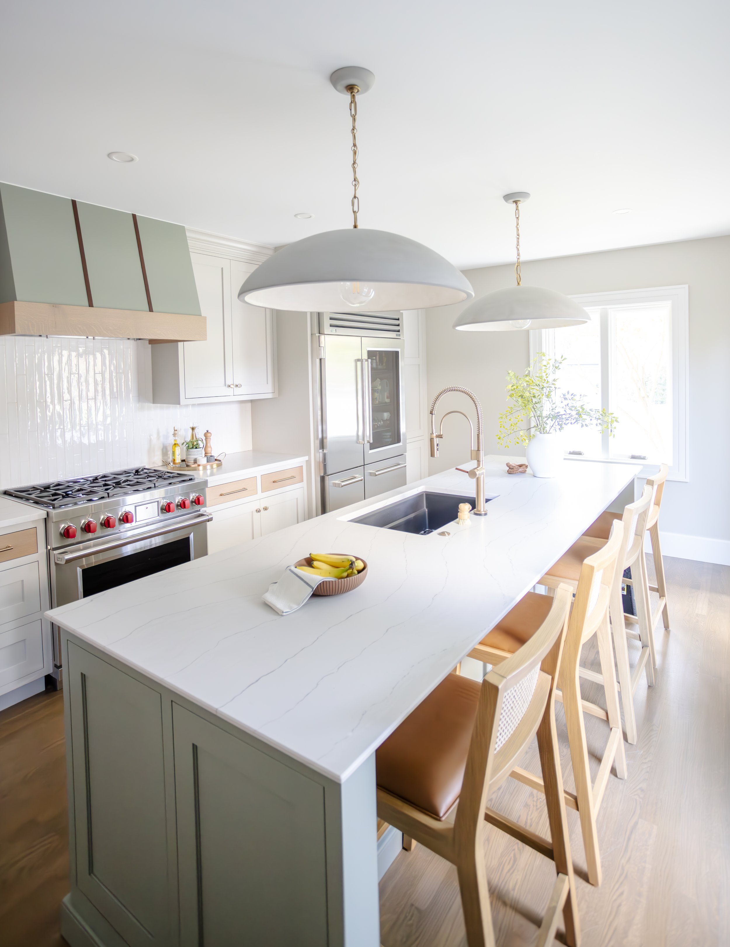 Bright kitchen with large quartz island, gas range with red knobs, soft green range hood, wicker chairs, earthy pendant lights, a vase with greenery, and a fruit bowl.