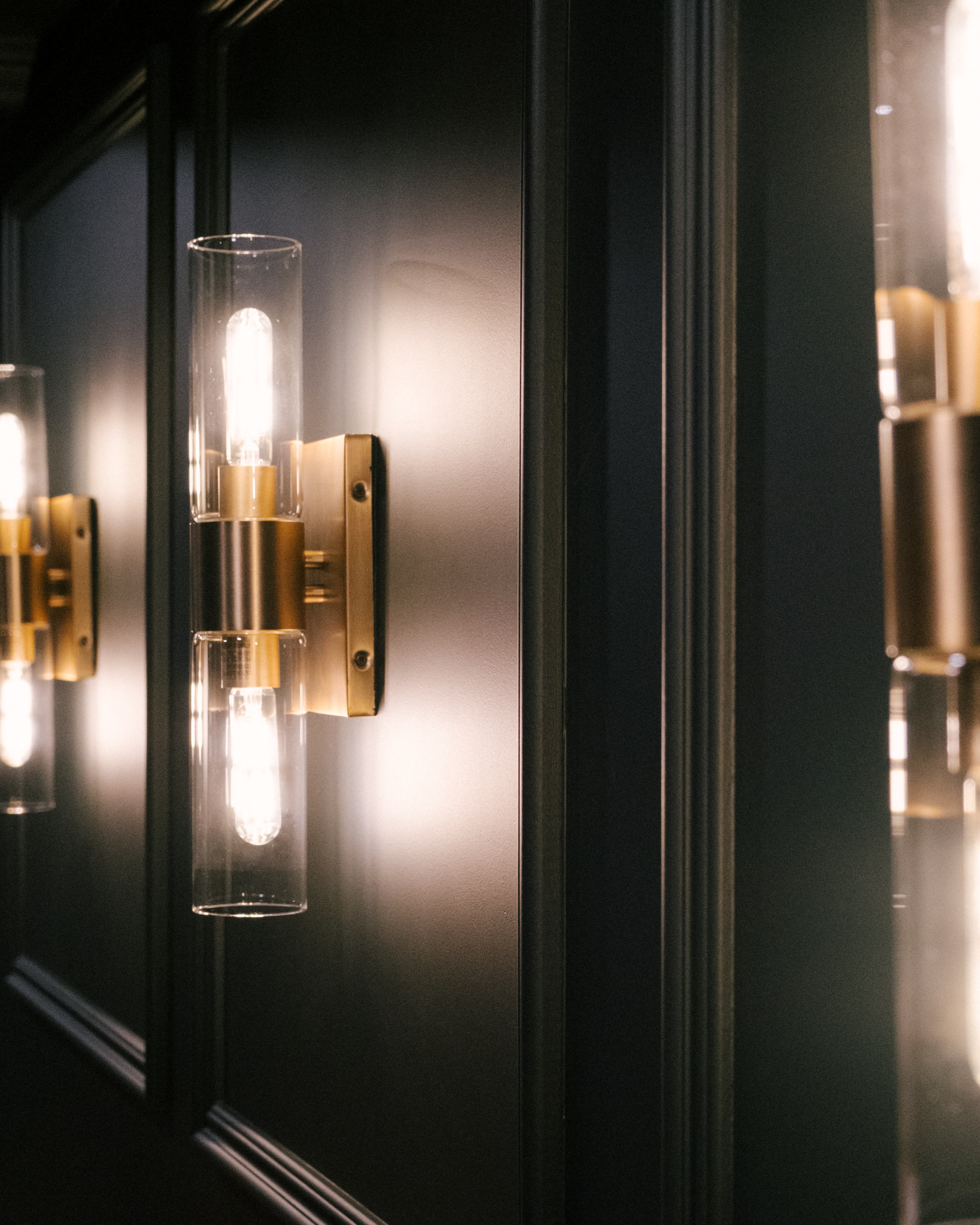 Modern wall sconces with clear glass shades illuminate a dark green, moody basement. The warm light creates a cozy and inviting ambiance.