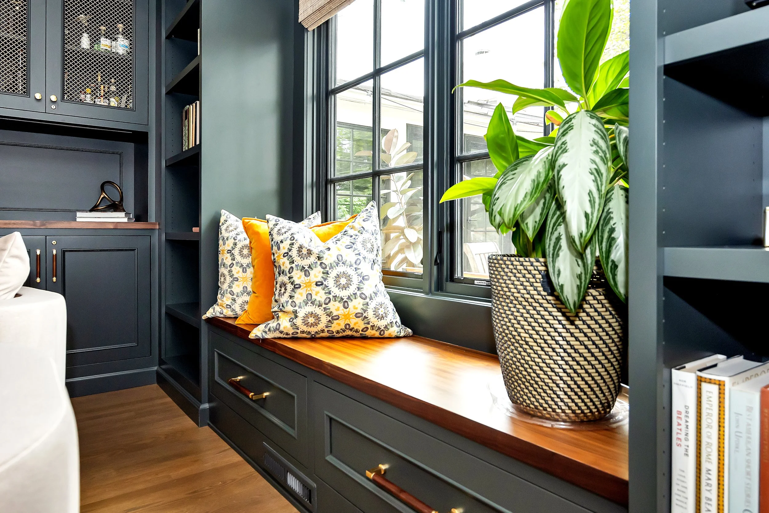 Cozy nook with a wooden bench under large windows, adorned with floral and yellow pillows. Moody green integrated library shelves are present, along with books, plants, and warm natural light.