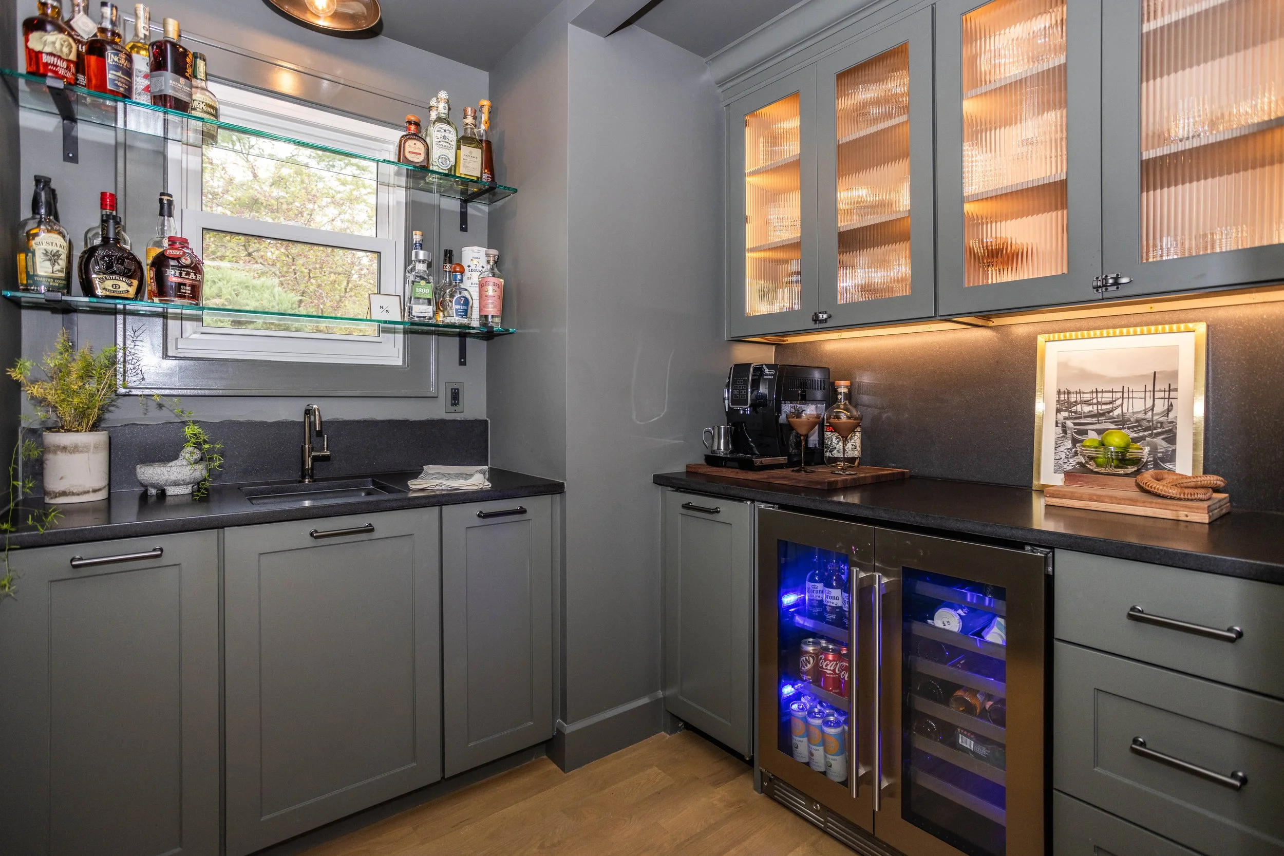 Stylish scullery and wet bar with modern gray cabinets, glass shelves showcasing liquor bottles, a window, and warm lighting. Sleek and inviting ambiance.