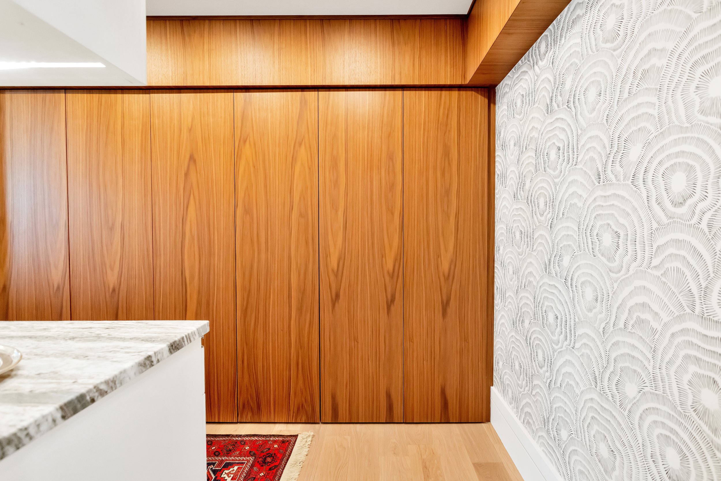 Modern interior with warm wood paneling and white textured wallpaper. A quartz countertop and part of a red rug peek into the frame, creating a cozy feel.