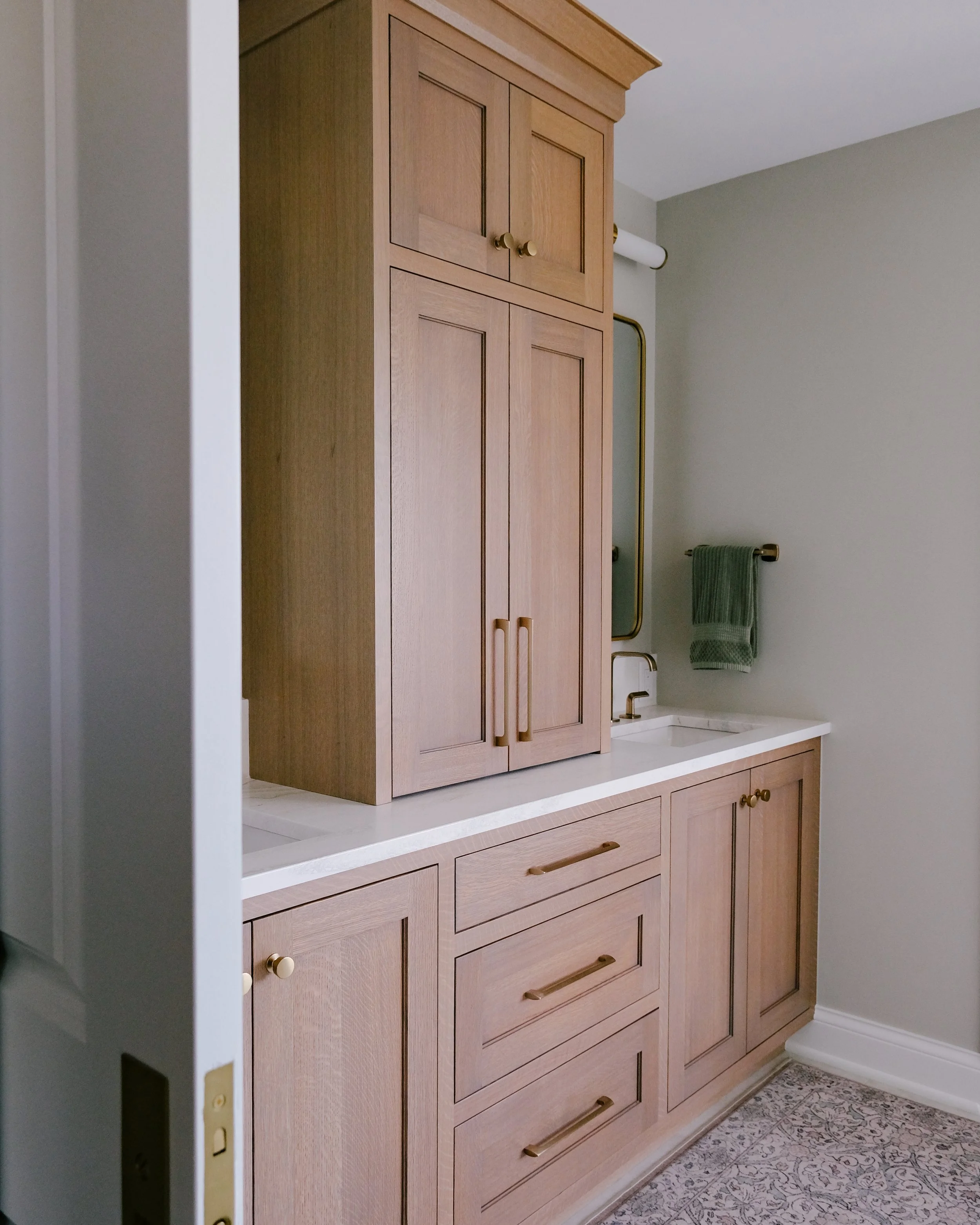 Modern bathroom with White Oak vanity and  cabinetry featuring honey bronze handles, a Cambria quartz countertop, luxe gold Brizo Allaria faucets, green towel, and a gold-rimmed wall-mounted mirror.