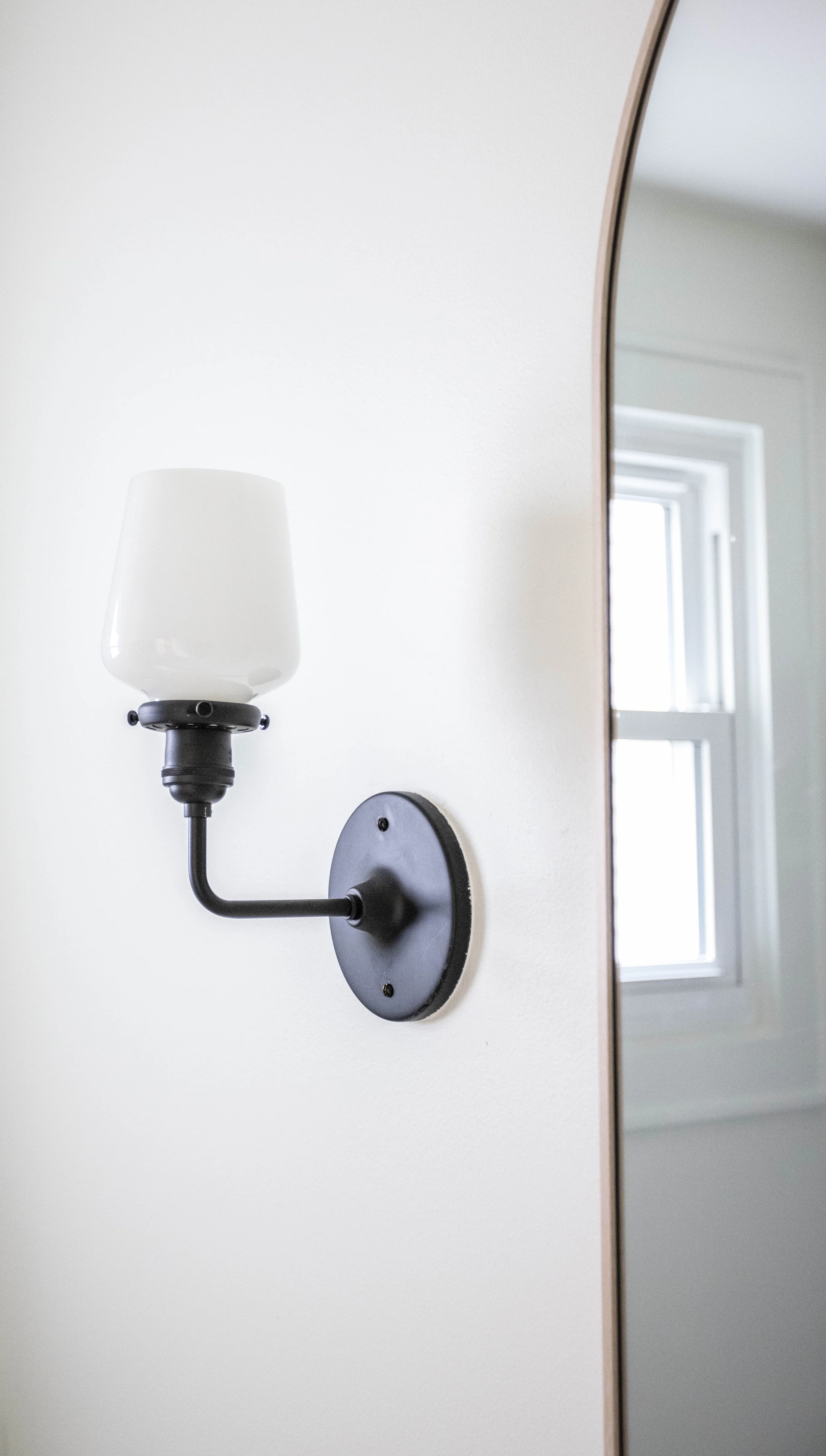 Wall-mounted black sconce with white frosted glass shade on an off-white wall near an arched mirror, bright window light softens the minimalist scene.