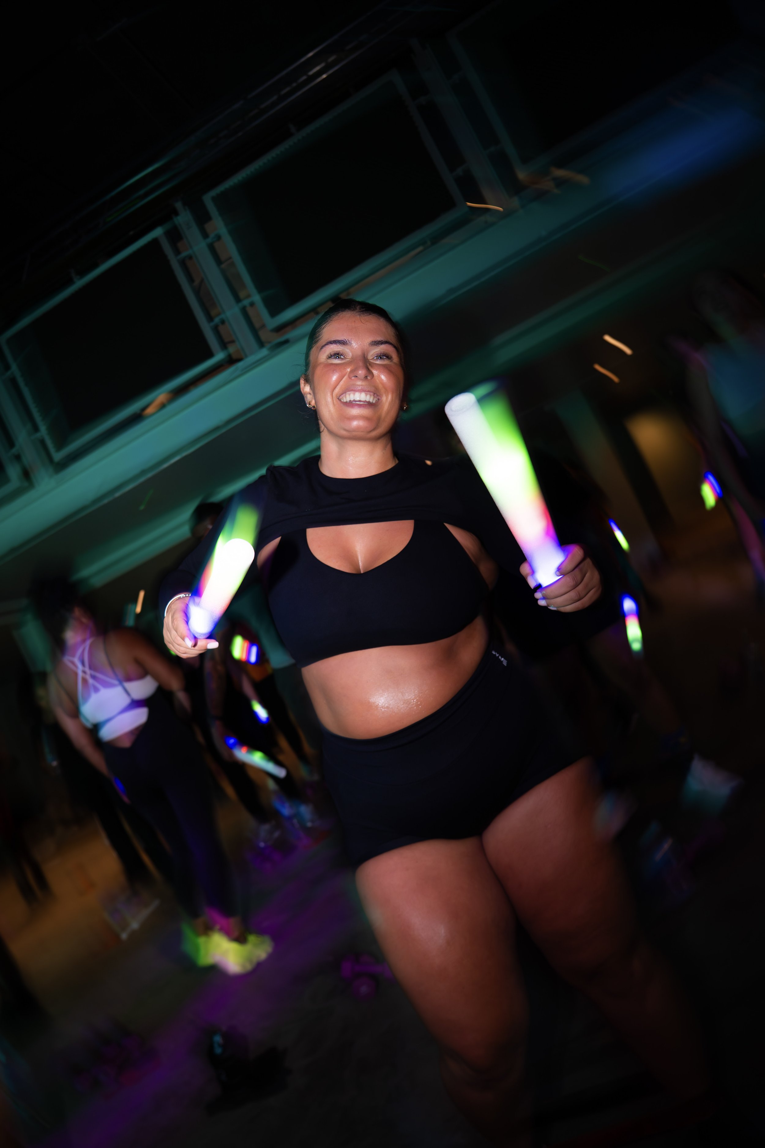 A woman smiling and holding glowing light sticks in a dark, lively party or club setting.