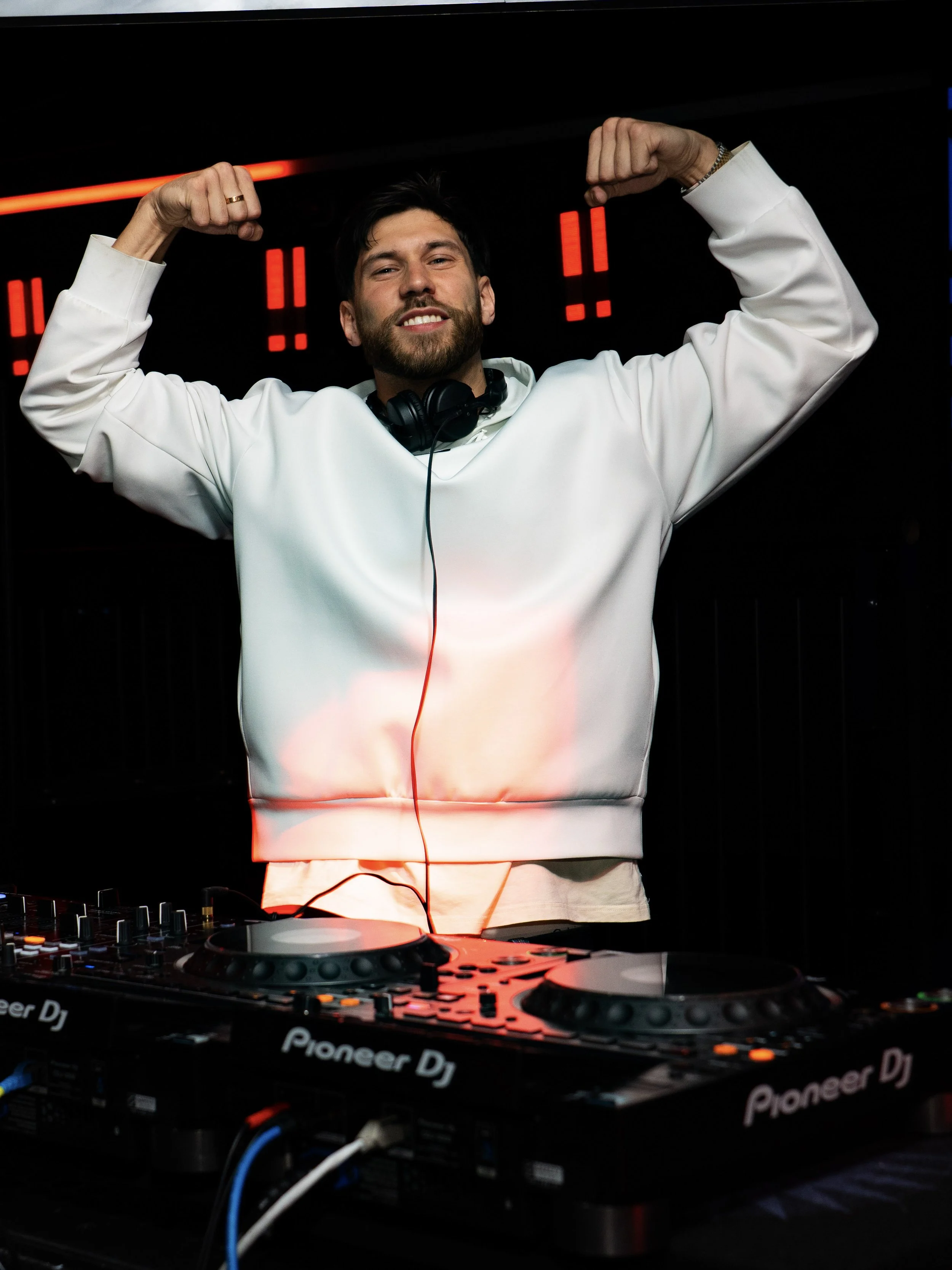 A man with a beard and dark hair, wearing a white sweatshirt, stands behind DJ equipment with Pioneer DJ logo, in a dark club, flexing his muscles in a victorious pose with a smile.