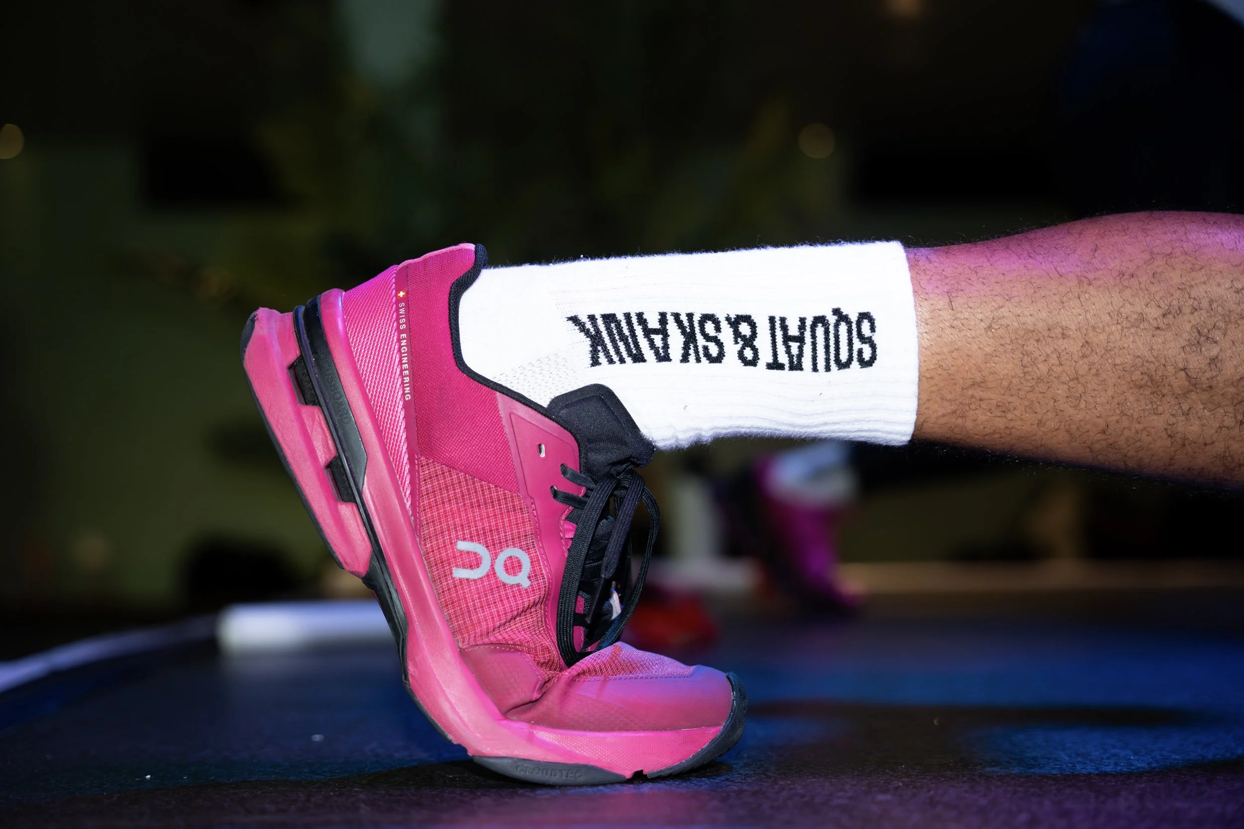 Close-up of a person's leg wearing a white sock with black text and a bright pink running shoe, with a blurred indoor background.