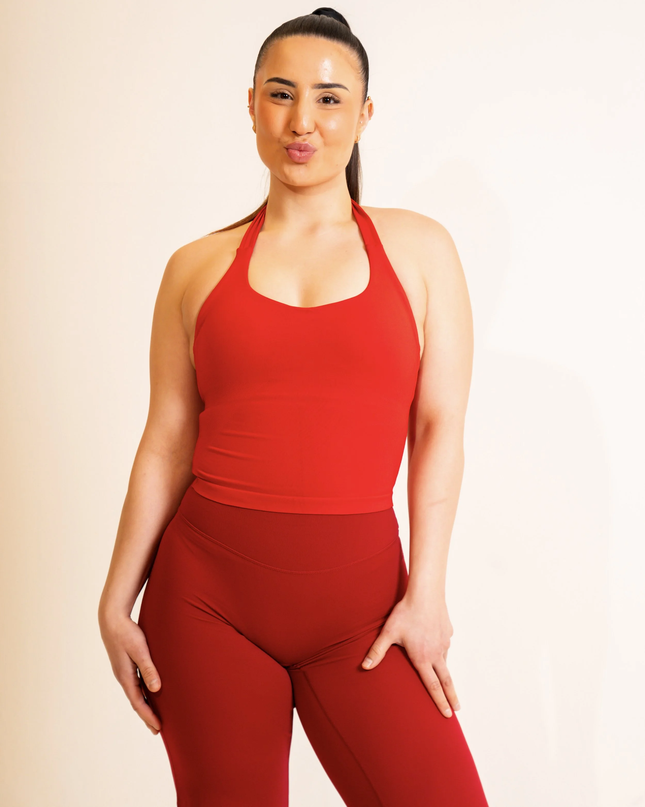 A woman wearing a red sports bra and matching red leggings, standing against a plain light background, making a playful kissing face.