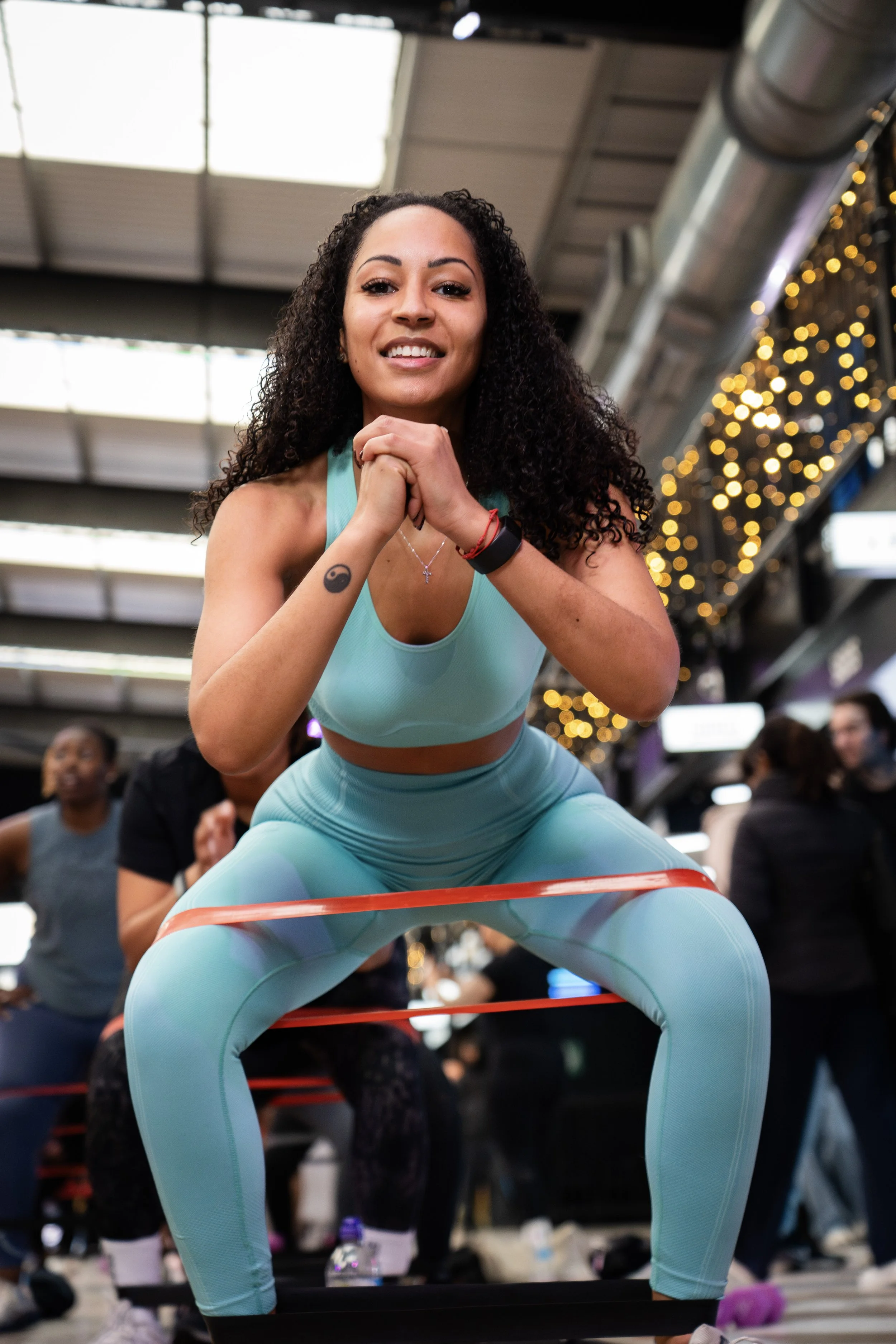 Woman in teal workout outfit using resistance bands in a gym with other people exercising in the background.