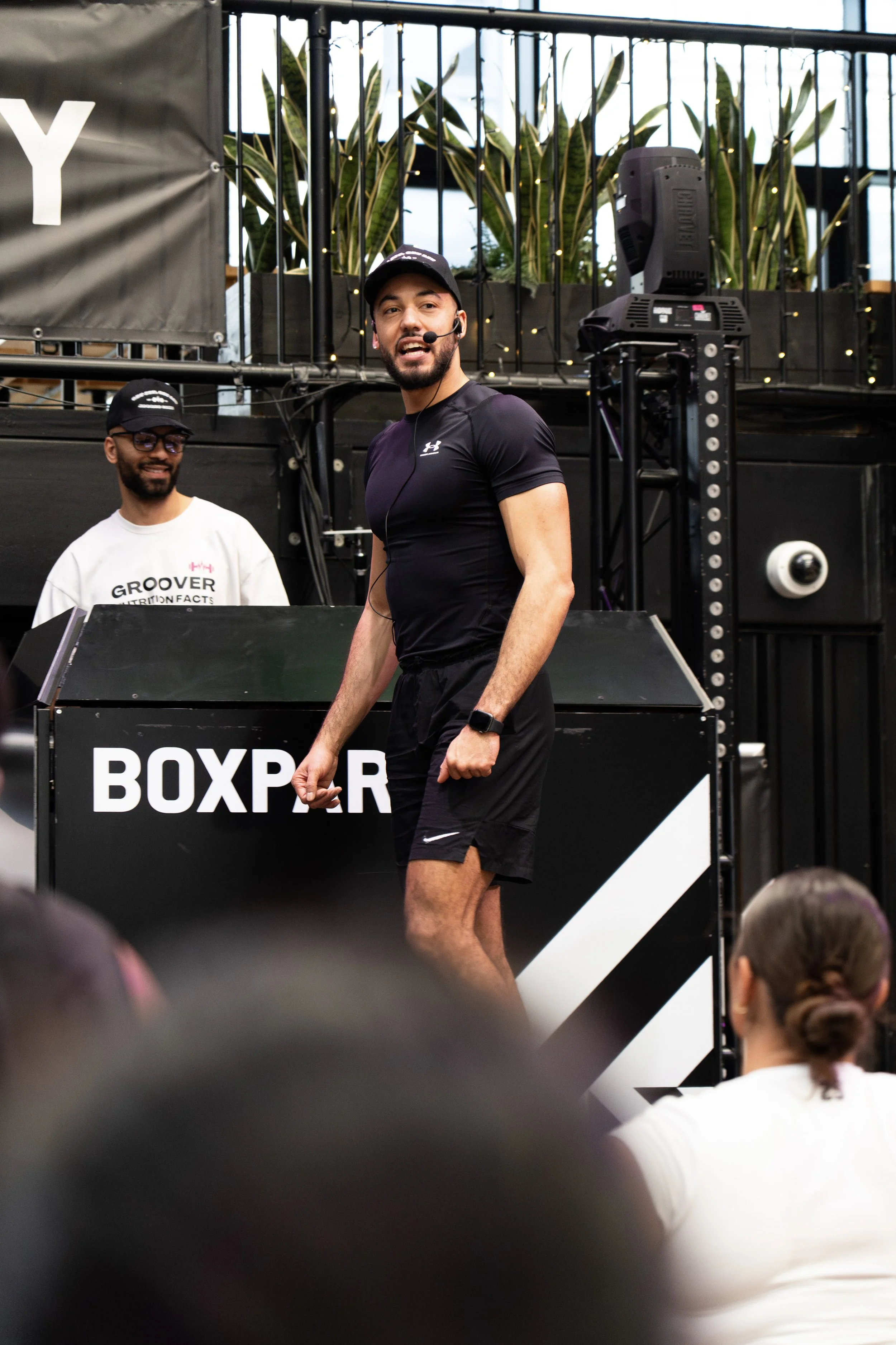 A fitness instructor with a microphone headset, wearing a black Under Armour shirt and black shorts, leading a workout class at a gym or fitness event. He is smiling and appears energetic. Another person with glasses and a white shirt stands behind a