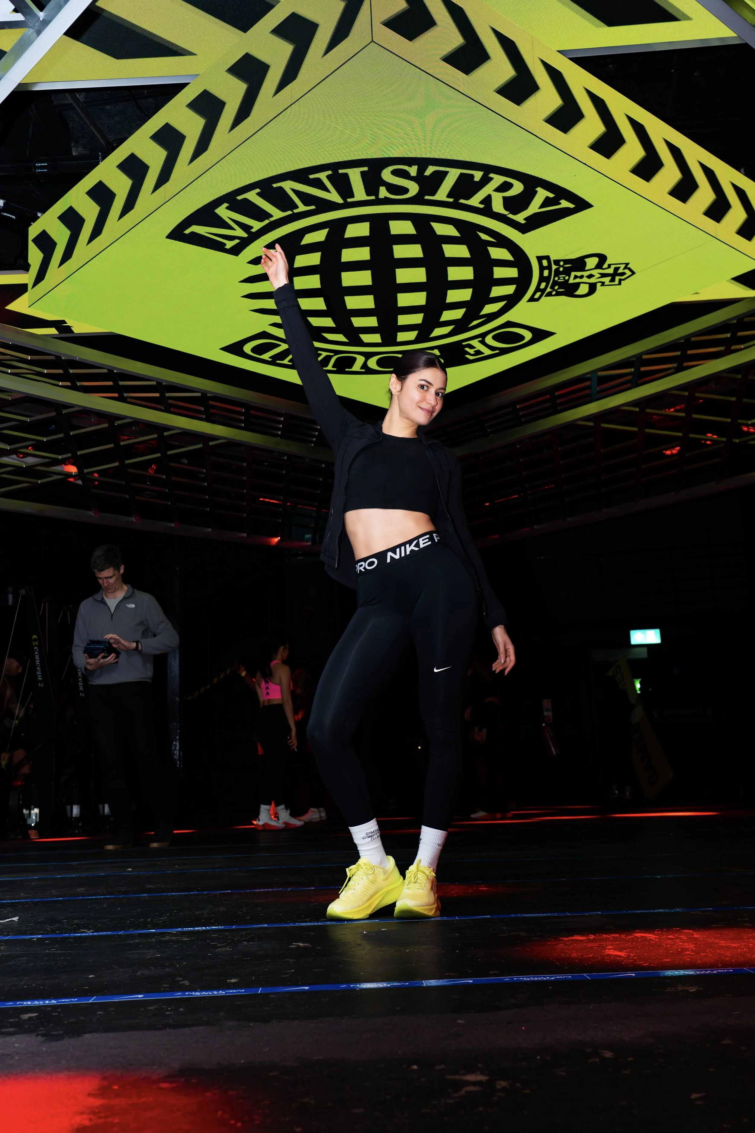 A woman in black athletic wear and yellow sneakers striking a pose at an indoor fitness event with a large Ministry of Sound logo overhead.
