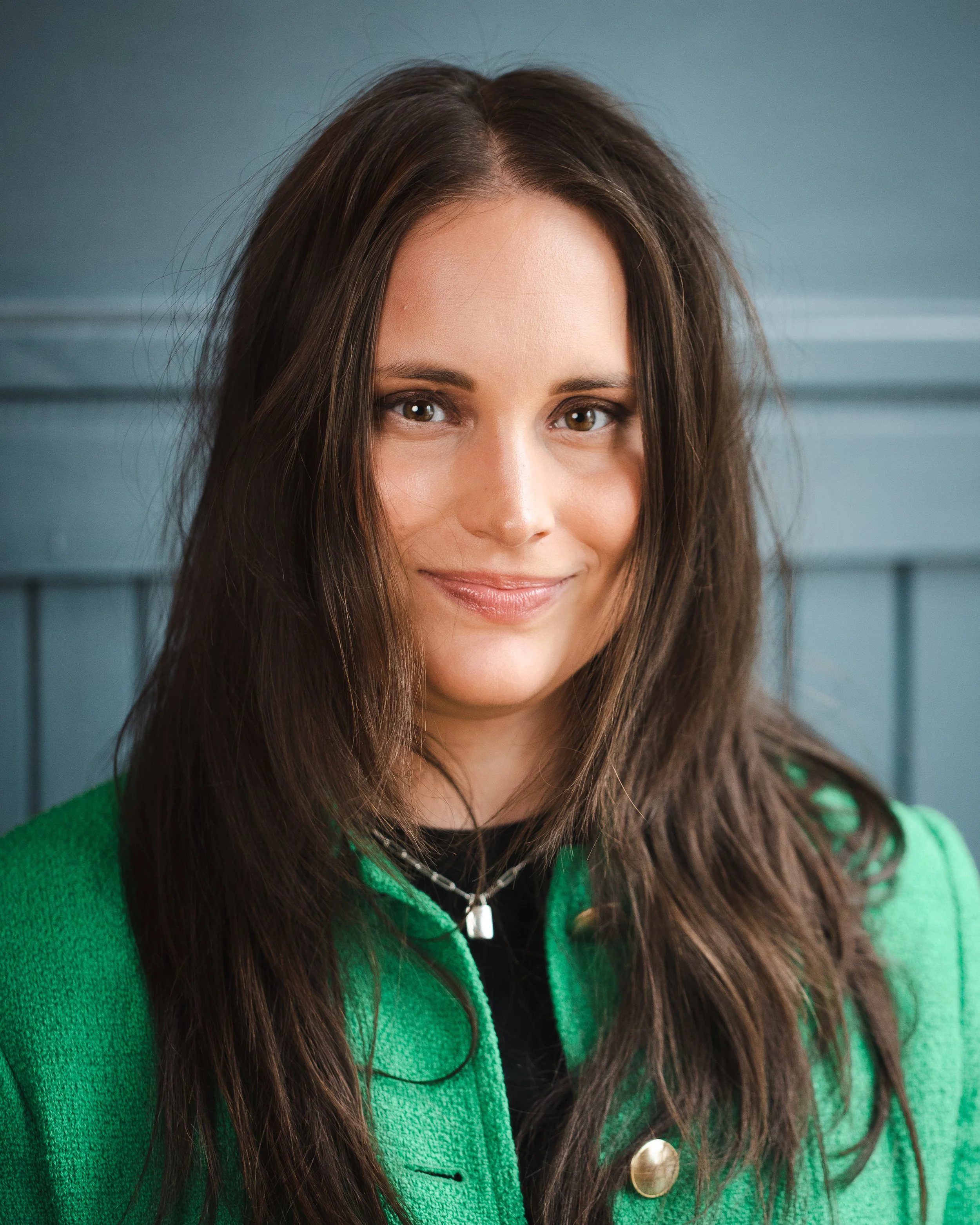 A woman with long brown hair, wearing a green jacket and silver necklace, smiling softly against a blue wooden background.